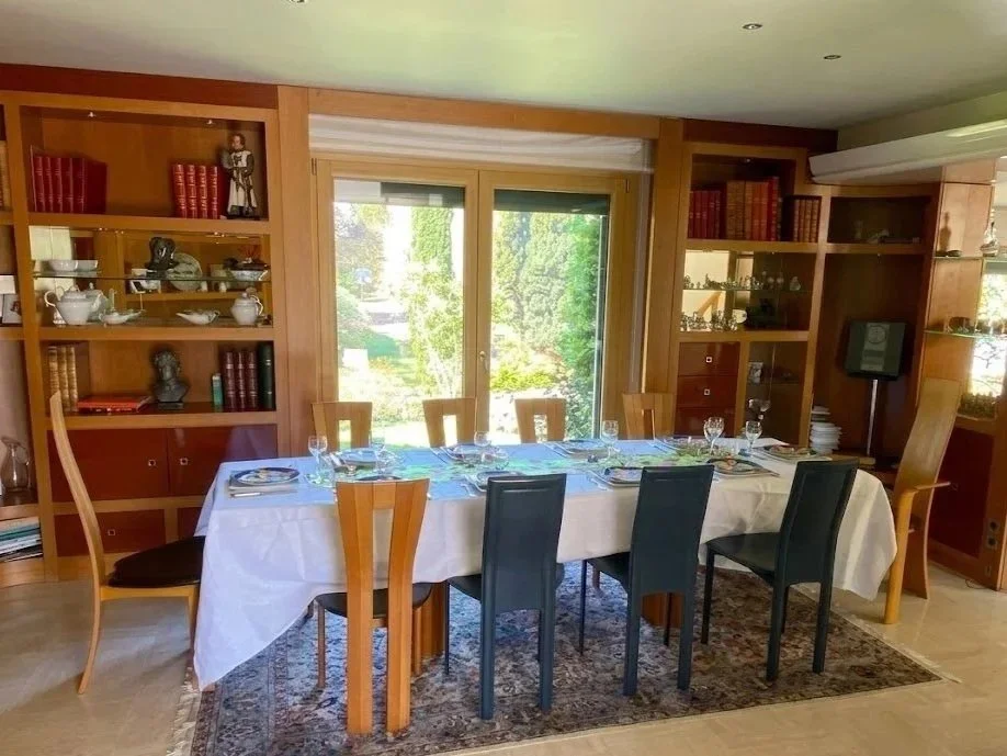 Salle à manger avec une grande table couverte d'une nappe blanche, entourée de plusieurs chaises, avec des étagères en bois remplies de livres et d'objets décoratifs derrière.