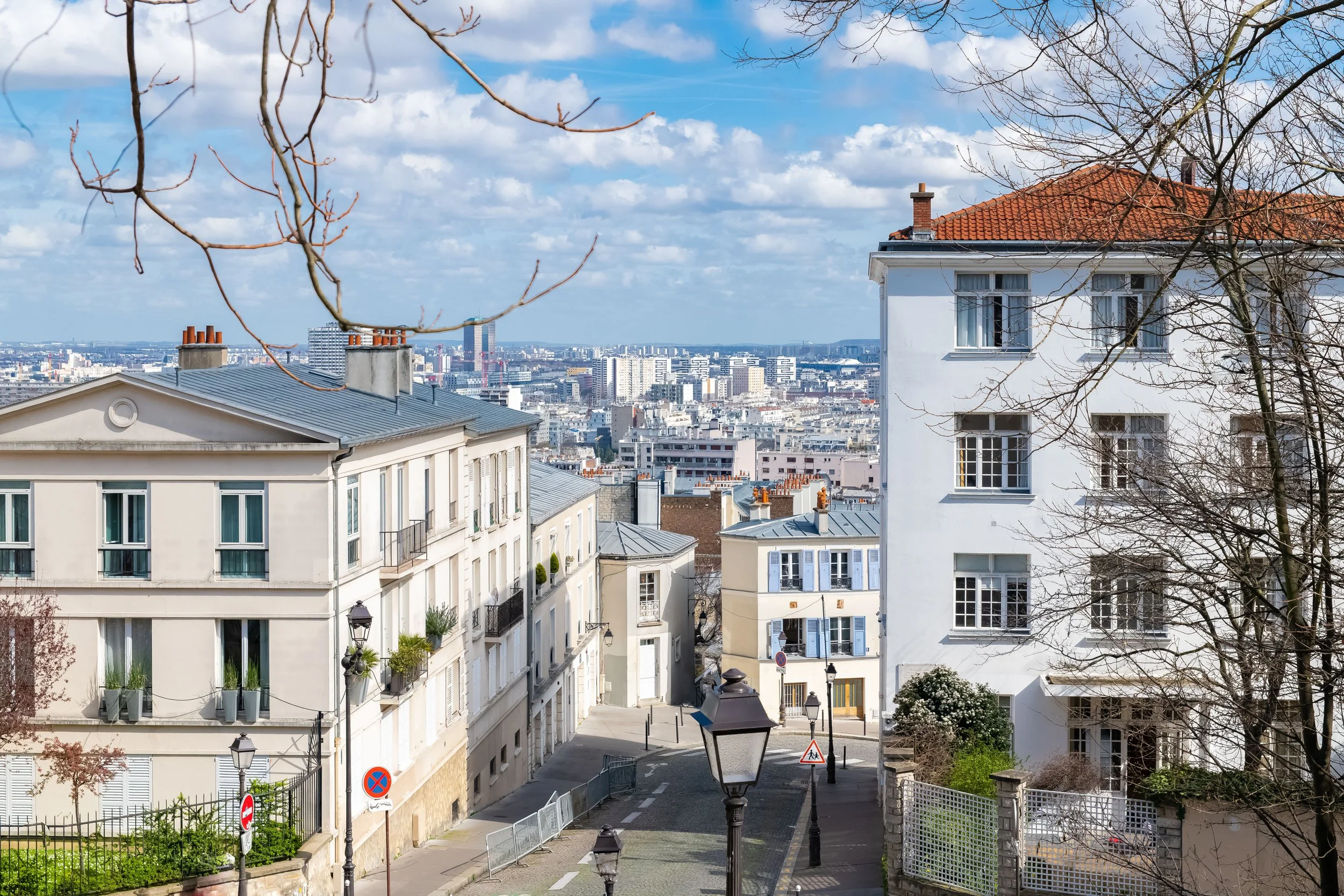 Vue de bâtiments résidentiels à Paris avec le ciel bleu et quelques nuages, un arbre sans feuilles et des lampadaires dans une rue en pente.