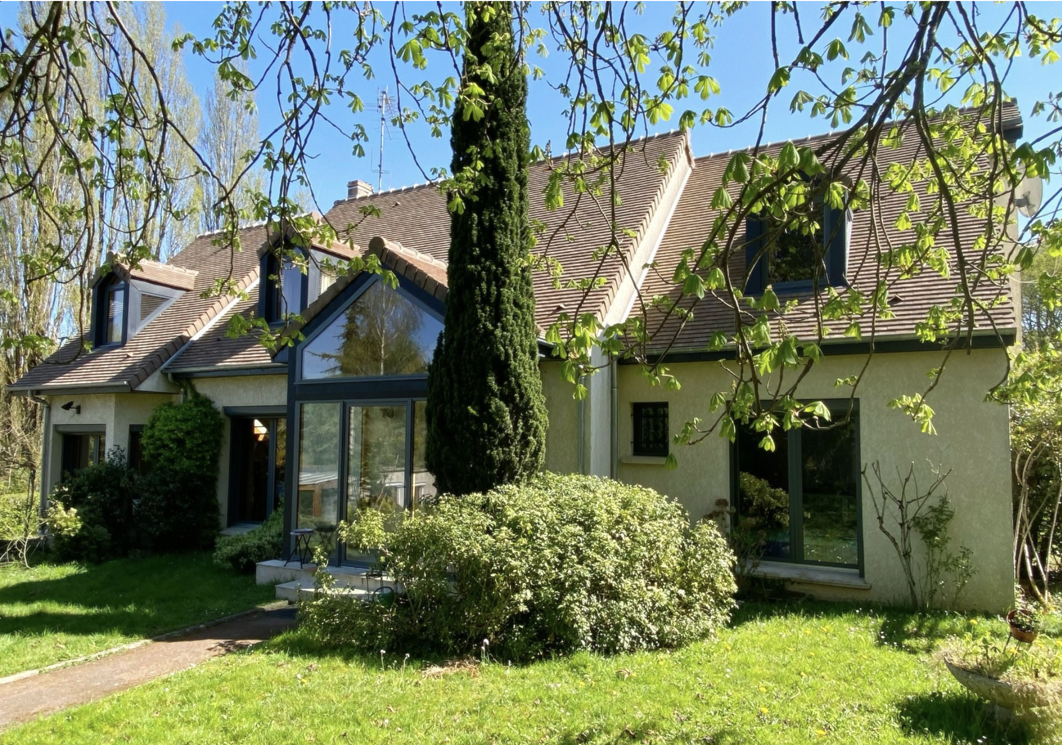 Maison avec plusieurs fenêtres en verre et toit en tuiles, entourée d'arbres et de végétation verte, sous un ciel bleu clair.