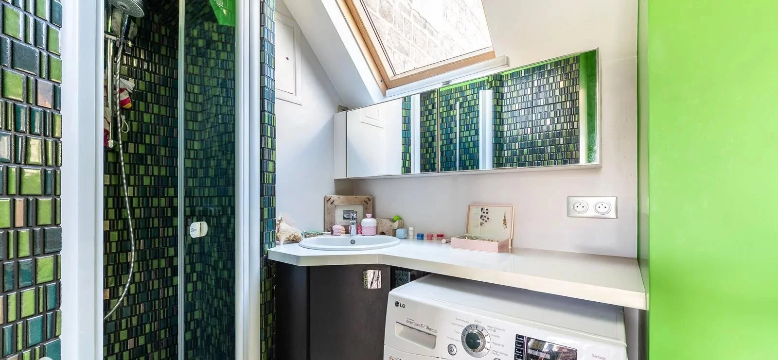 Petite salle de bain avec mur en carreaux verts et noirs, un lavabo avec des produits de toilette, un miroir, une fenêtre de toit laissant entrer la lumière, une machine à laver et une prise électrique.