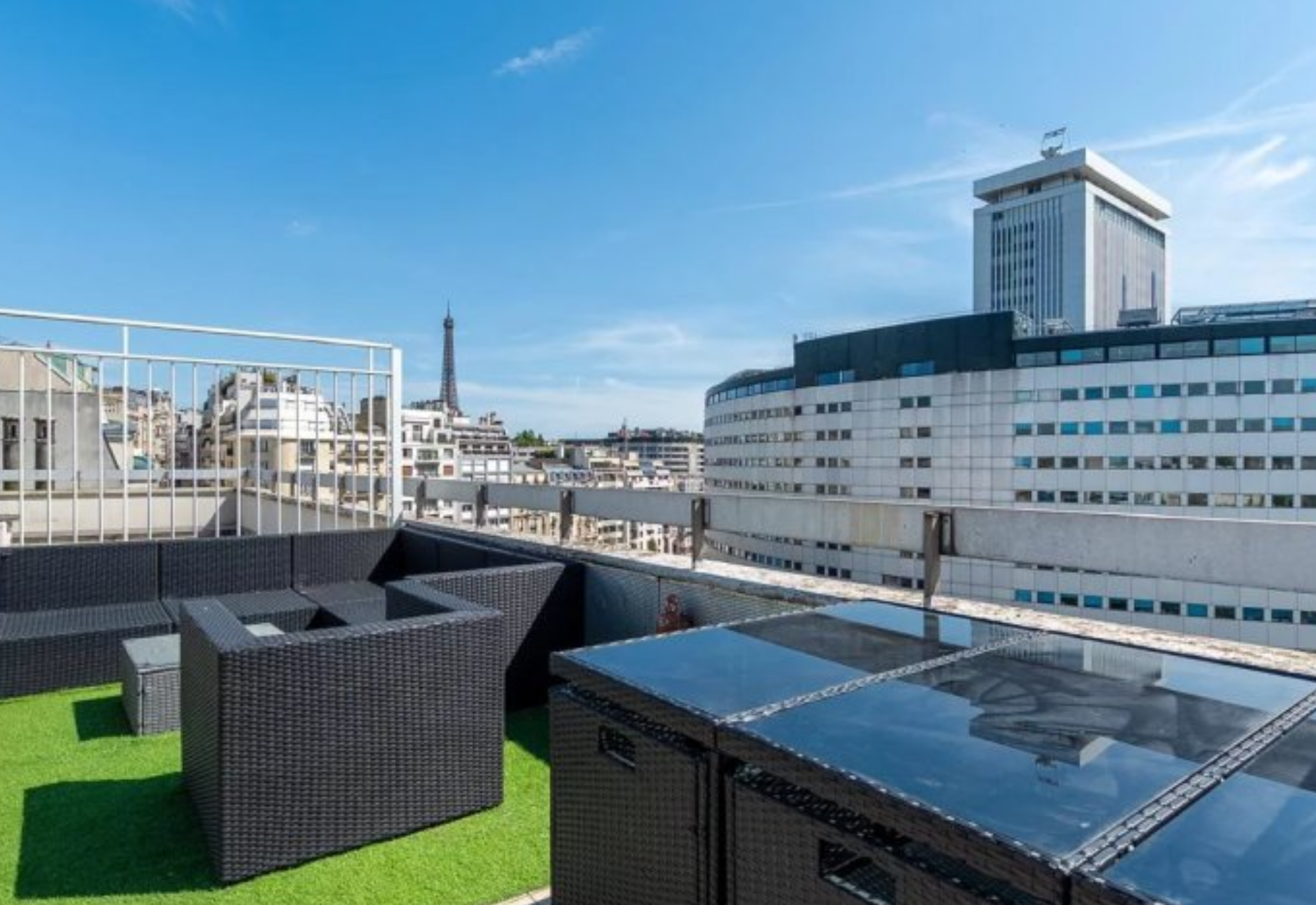 Terrasse urbaine avec mobilier en rotin noir et tapis vert, vue sur des bâtiments modernes et la Tour Eiffel au loin sous un ciel bleu clair.