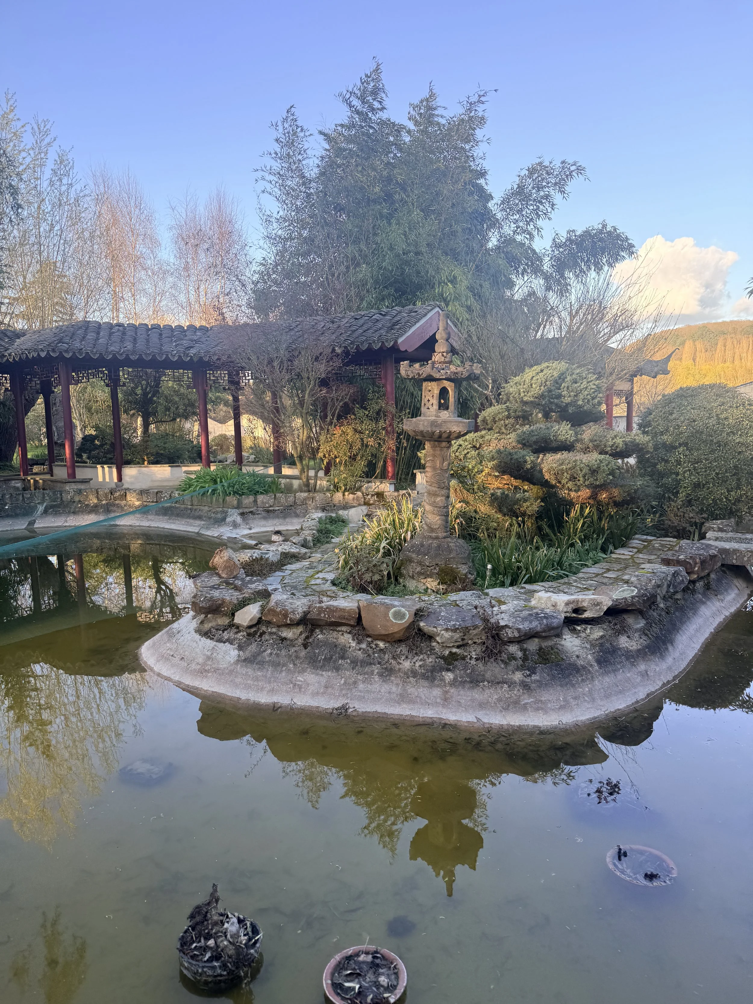 Un jardin japonais avec un étang, un pont en pierre, une lanterne traditionnelle en pierre, des arbres et des buissons, sous un ciel bleu avec quelques nuages.