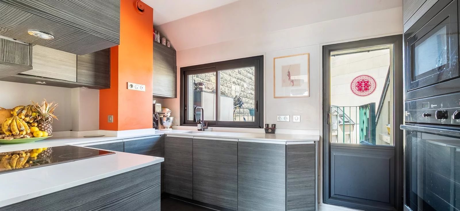 Cuisine moderne avec fenêtres avec vue sur un mur en pierre, étagères en bois foncé, comptoir blanc, fruits, décoration murale et porte-fenêtre donnant sur une terrasse extérieure.
