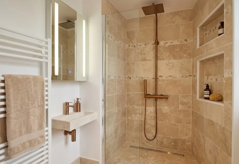 Salle de bain moderne avec douche à l'italienne en carrelage beige, lavabo blanc, miroir avec éclairage, et étagères encastrées avec produits de soins.