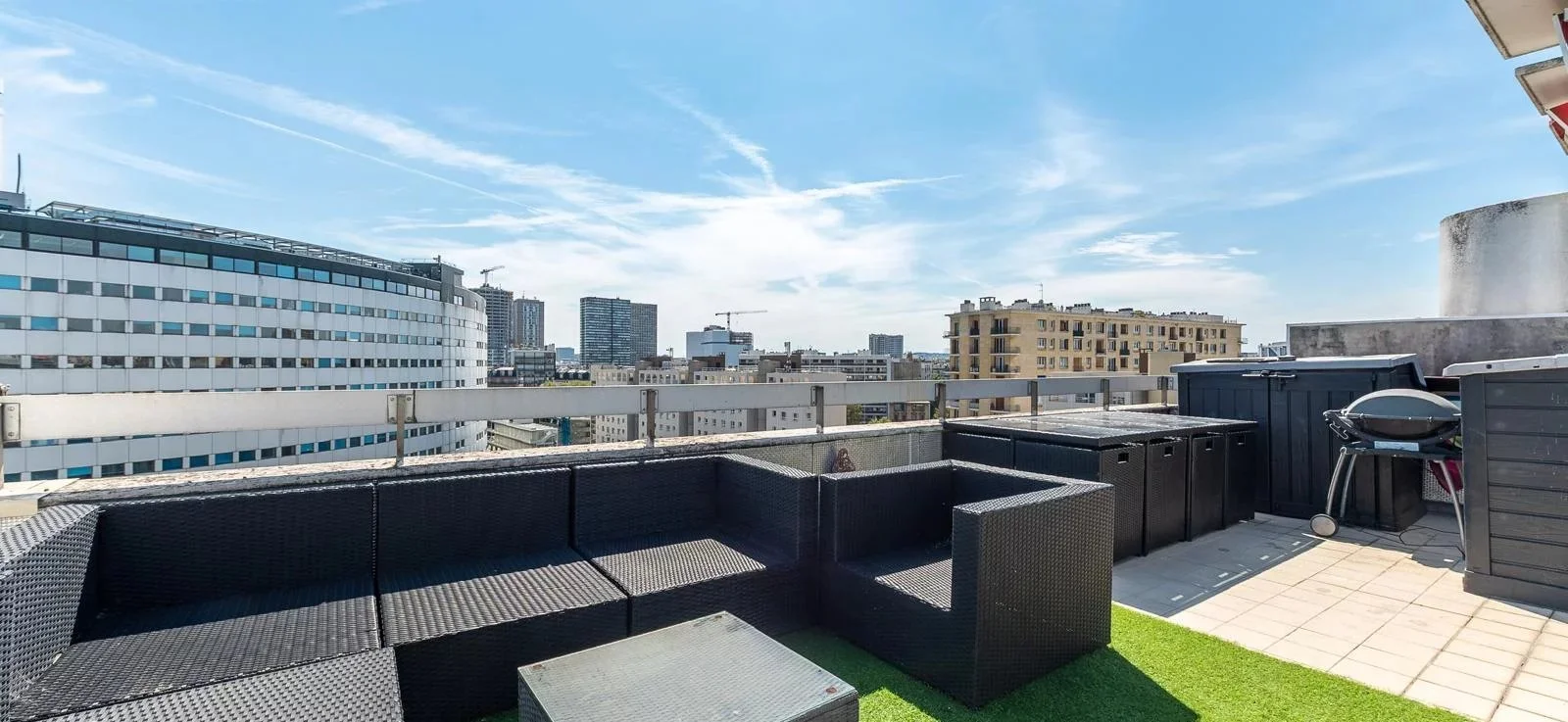 Terrasse urbaine avec mobilier noir, barbecue, vue sur des bâtiments modernes, ciel ensoleillé avec quelques nuages.