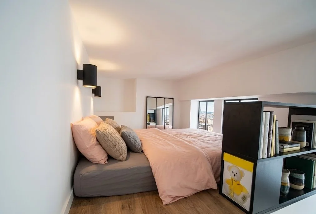 Chambre moderne avec un lit face à une fenêtre, bibliothèque noire à côté, murs blancs, sol en bois, et éclairage mural