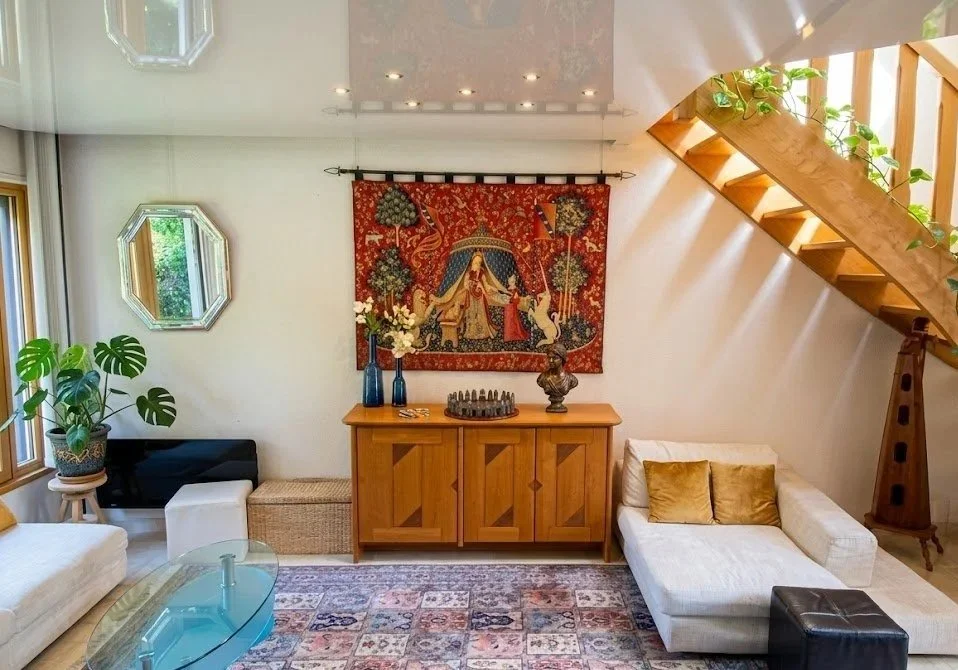 Salon lumineux avec canapé blanc, tapis, étagère en bois, décor de tapisserie colorée, plantes, miroirs octogonaux, et escalier en bois avec plantes vertes.