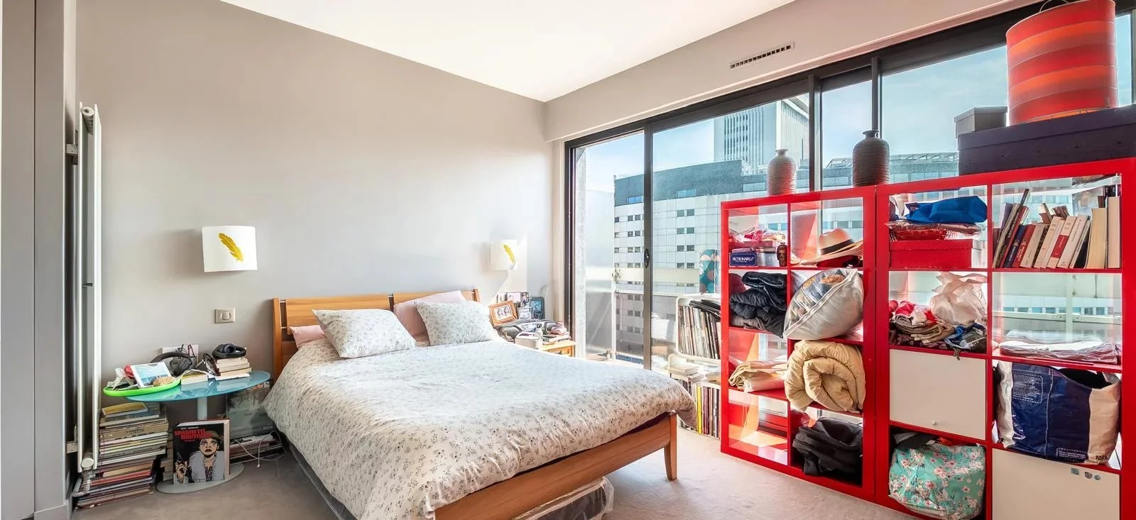Chambre avec un lit, deux lampes murales, une petite table de nuit avec des livres et objets, un gros meuble rouge avec des vêtements, sacs et livres, grandes fenêtres avec vue sur des immeubles modernes.