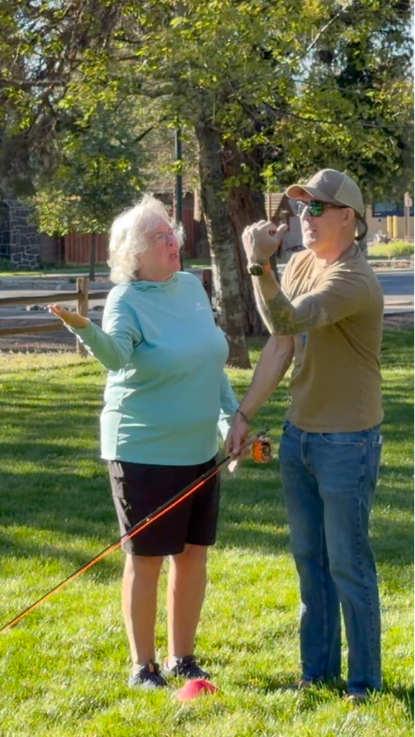 A woman in a light blue long-sleeve shirt and black shorts holding a golf club stands on a grassy field, talking to a man in a beige cap, sunglasses, and khaki shirt, who is pointing upwards. The scene is outdoors with trees and a street in the background.