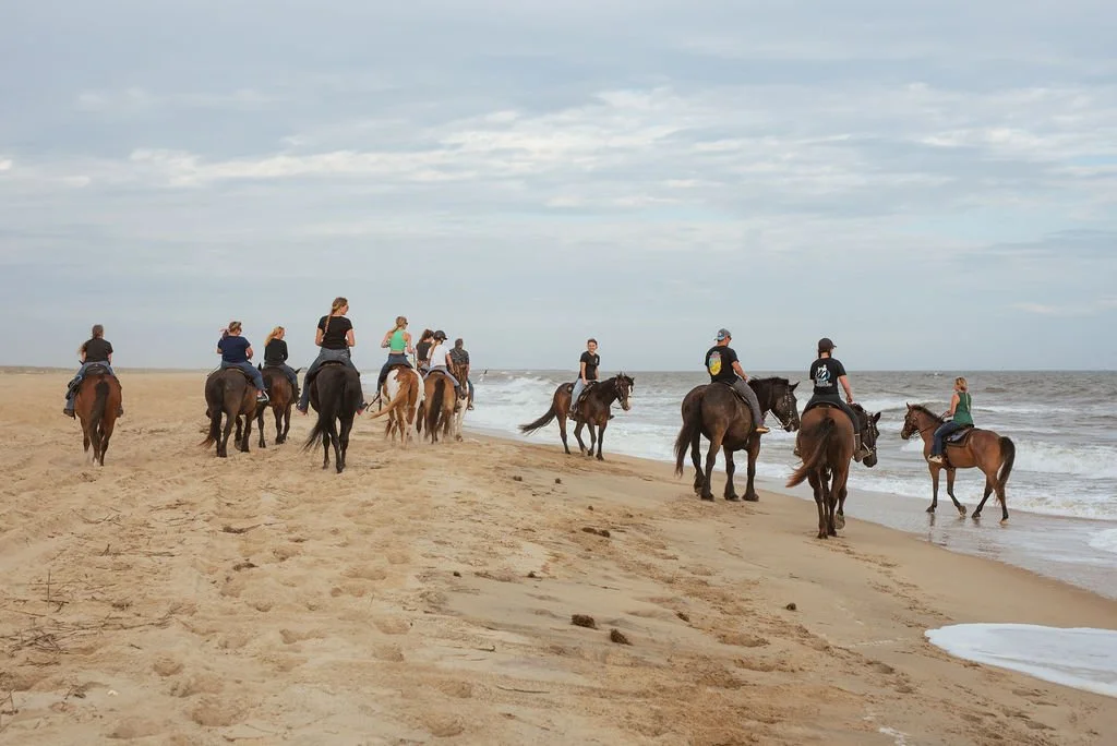 People horseback riding on the beach near the water with a cloudy sky.