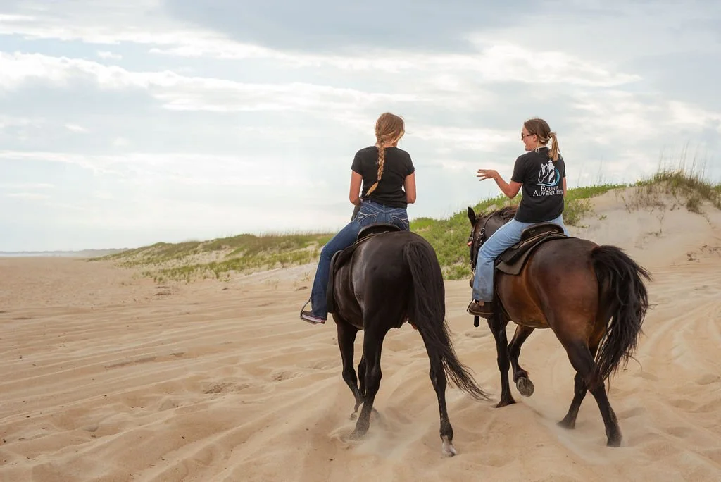 Two women riding horses on a sandy beach with dunes and grass, under a cloudy sky.