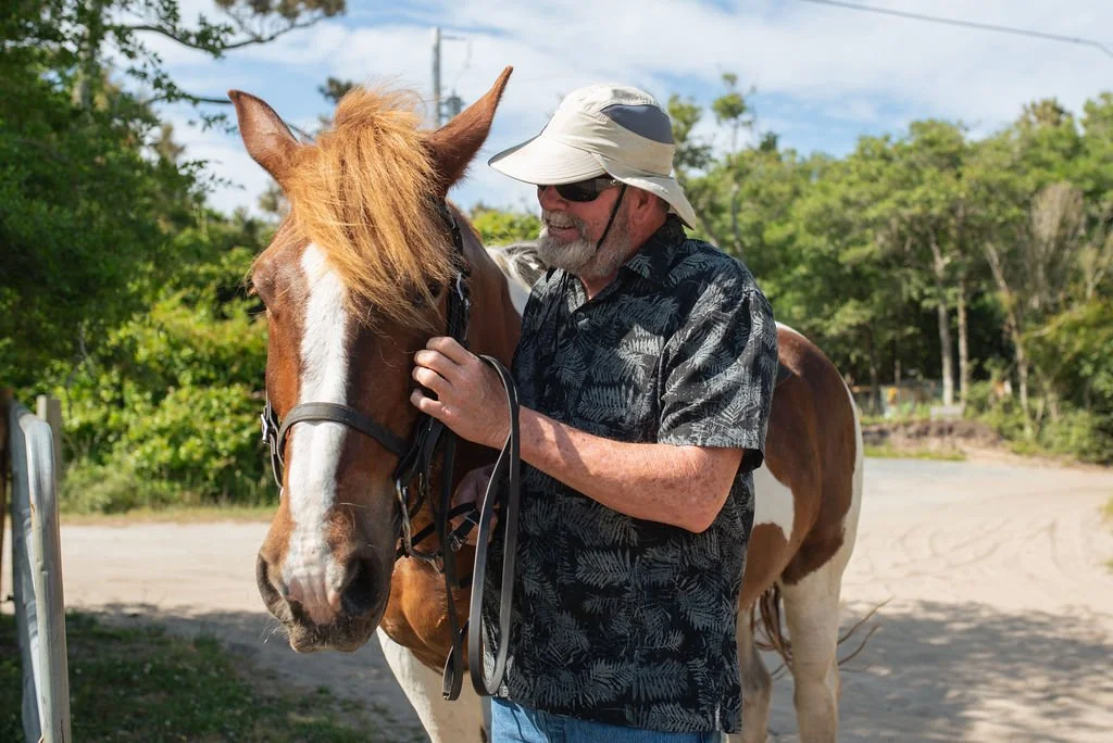 A man wearing a hat and sunglasses standing next to a brown and white horse, holding its bridle, outdoors in a rural setting.