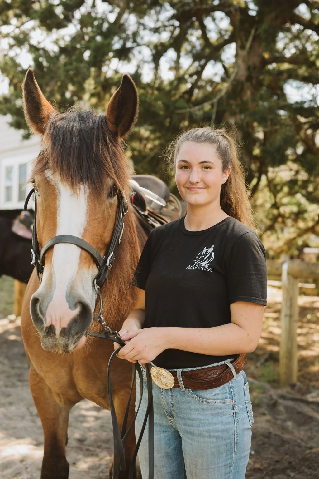 A young woman stands outside holding the reins of a brown and white horse, smiling at the camera. She is wearing a black t-shirt with a logo and light blue jeans.