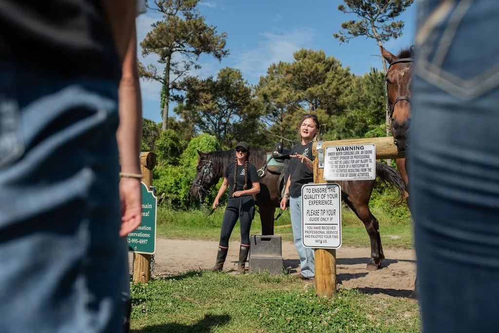 Two women standing with horses near informational signs outdoors, trees and blue sky visible.