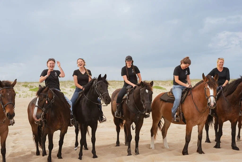 Five women riding horses on a sandy beach, some laughing and smiling, wearing black shirts.