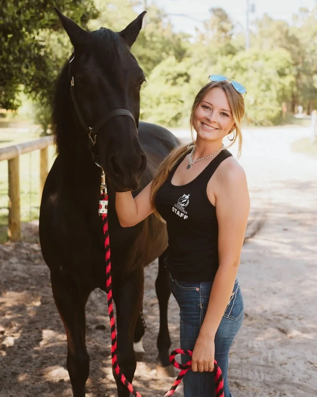 A young woman with blonde hair in a ponytail, wearing sunglasses, a black tank top, and jeans, standing outdoors on a dirt path with a smile, holding the lead rope of a large black horse beside her.