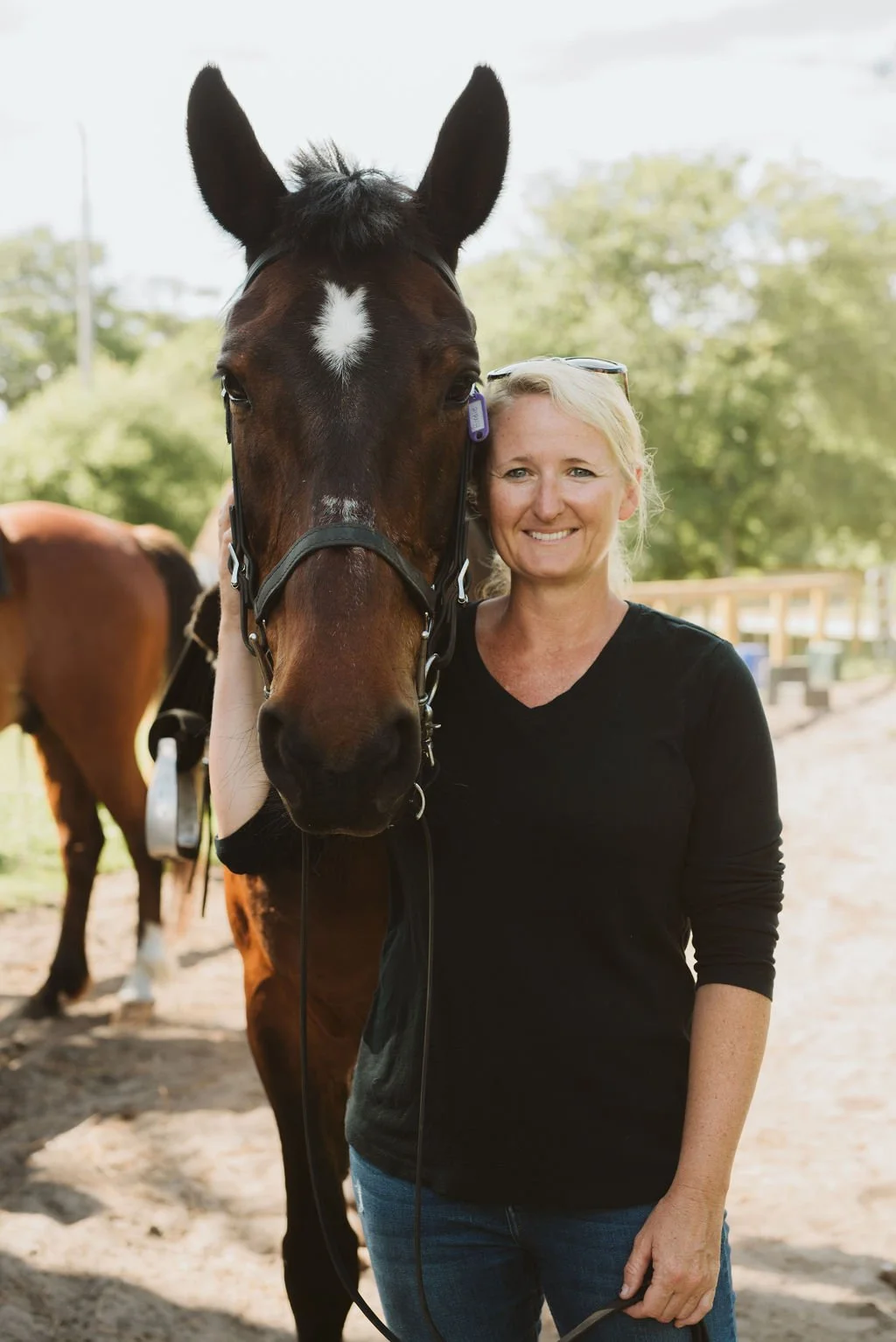 A woman smiling next to a dark brown horse with a white star on its forehead at an outdoor stable.