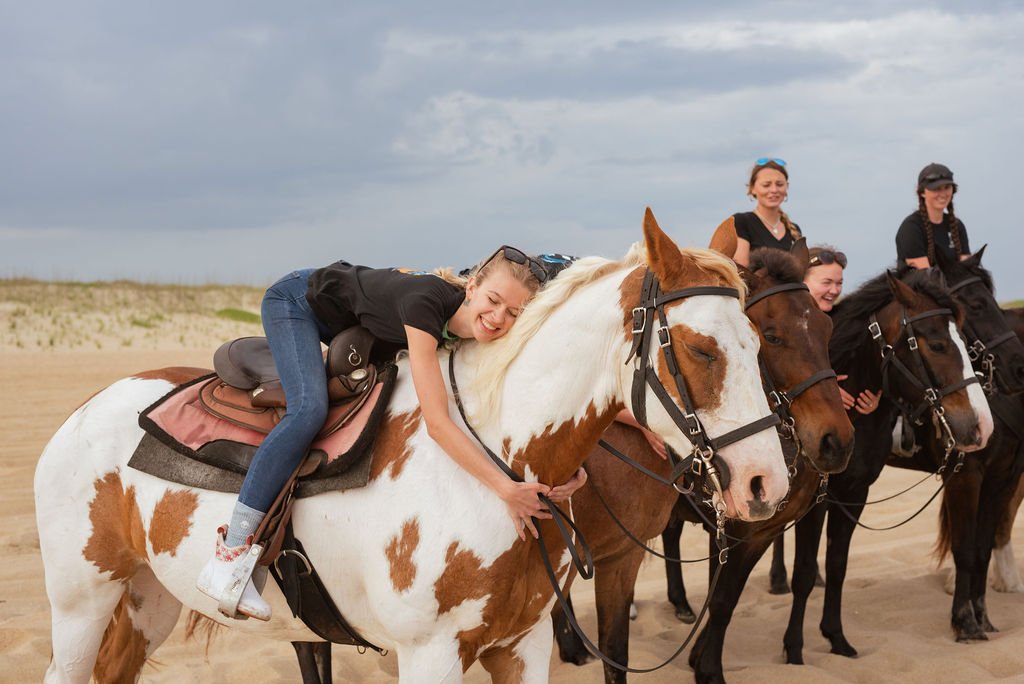 A young girl lying on a white and brown horse, smiling, with her arms around its neck on a sandy beach, while other people on horseback are in the background under a cloudy sky.