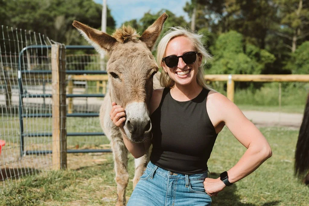 A woman with blonde hair, wearing sunglasses, a black sleeveless top, and jeans, smiling next to a donkey in a fenced outdoor area.