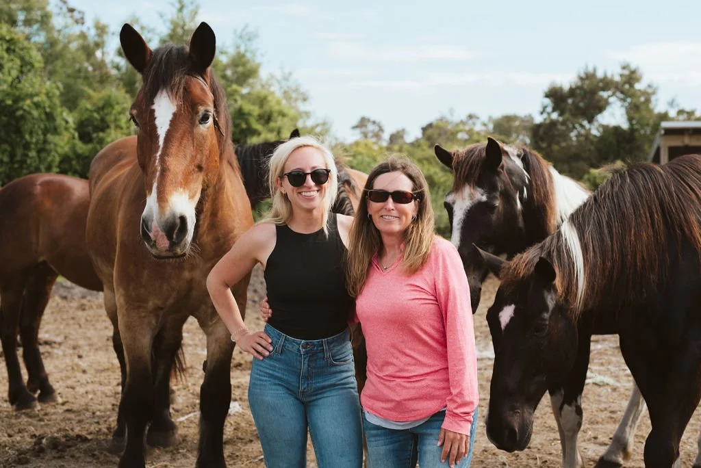 Two women standing close together in front of several horses outdoors, with greenery and trees in the background.
