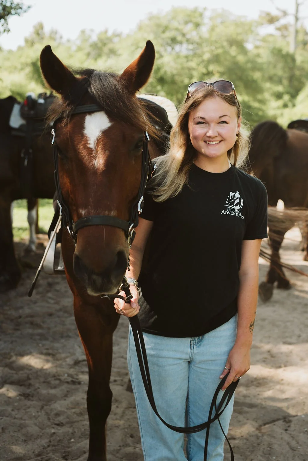 A young woman in a black t-shirt holding a brown horse on a lead rope, standing outdoors in a green, wooded area.