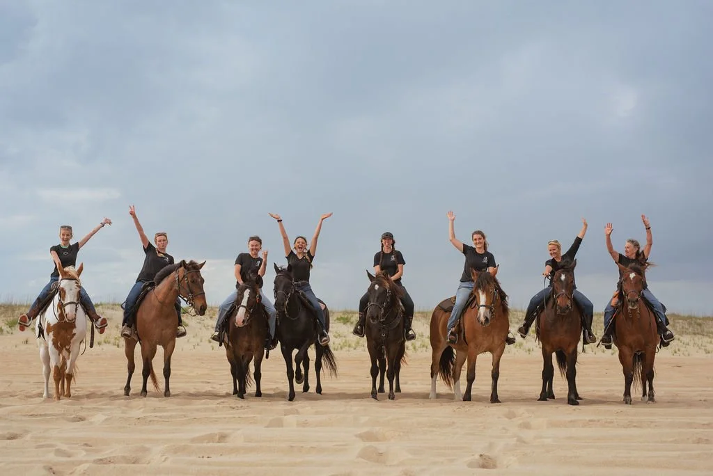 Eight women riding horses on a sandy beach with overcast sky, all waving and smiling.
