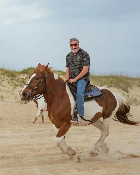 A man with gray hair and sunglasses riding a pinto horse on a sandy beach, smiling with a cloudy sky in the background.