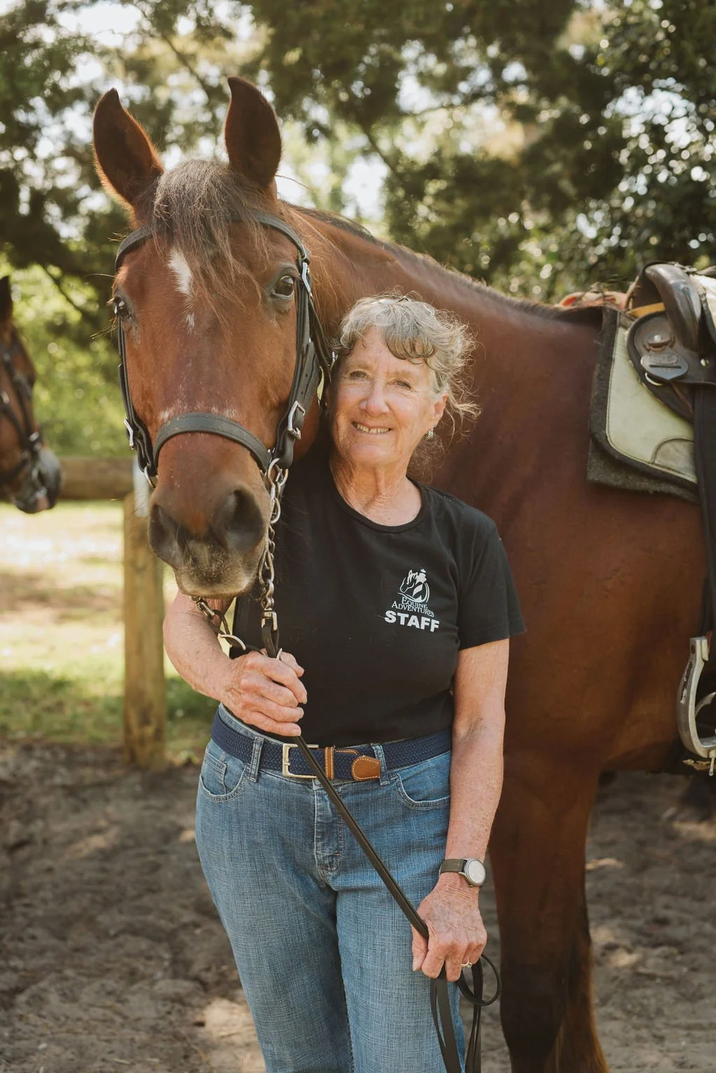 A woman standing outdoors with a brown horse, smiling and holding its bridle, wearing a black t-shirt and jeans.