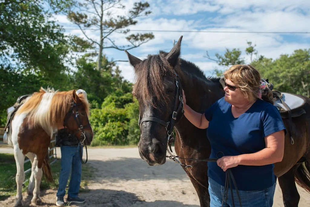 A woman in a blue shirt and sunglasses petting a black horse, with another person in the background holding a brown and white horse in a sunny outdoor setting with trees and a blue sky.
