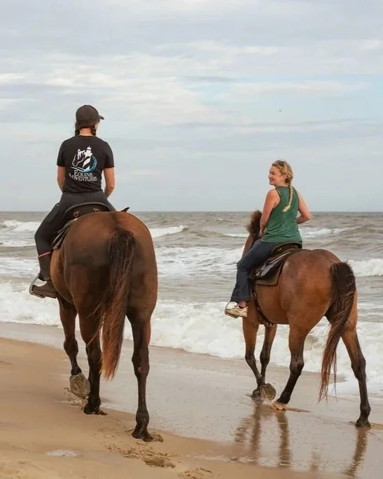 Two women riding horses along the beach with ocean waves in the background.