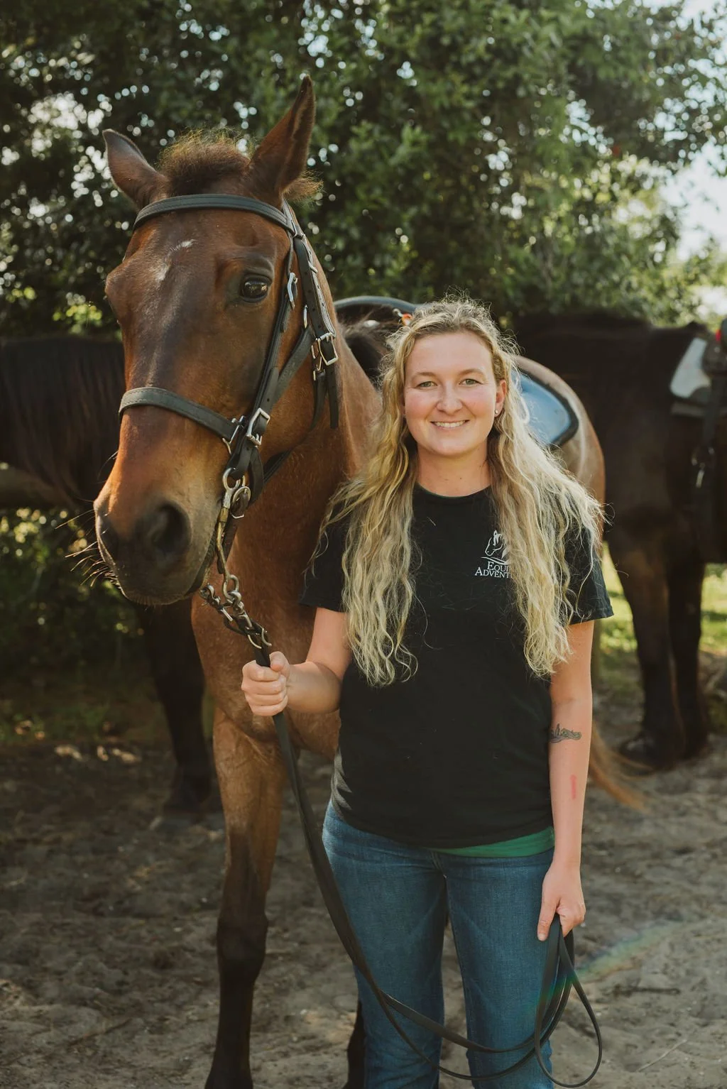A smiling woman with long blonde hair standing outdoors, holding the reins of a brown horse with a black saddle, in front of green trees and a few parked cars in the background.