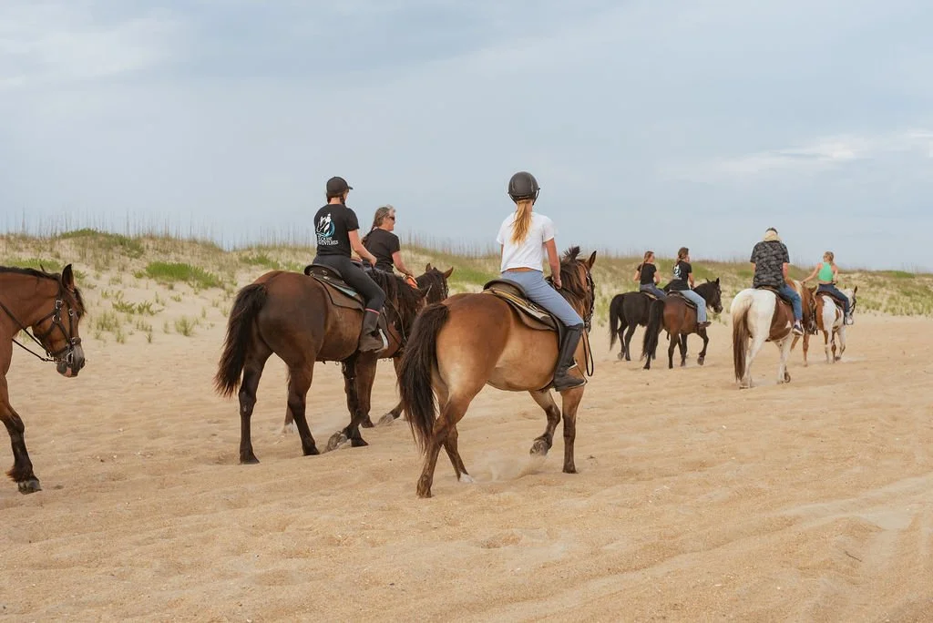 Group of people horseback riding on a sandy beach with grassy dunes in the background.