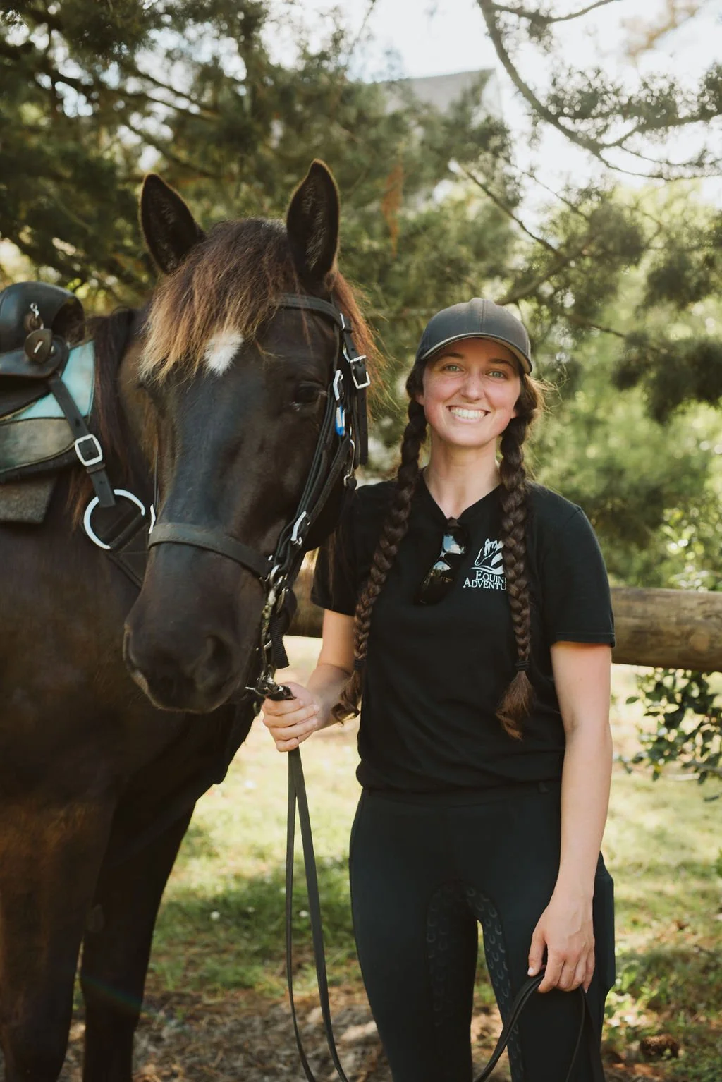 A woman with two braids and a black cap smiling while standing next to a brown horse outdoors, holding the horse's leash. The woman is wearing a black polo shirt and black pants, and the horse has a saddle and bridle. There are trees and a wooden fence in the background.