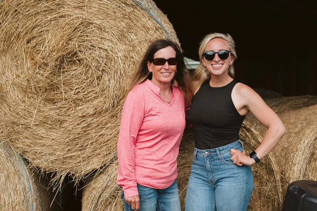 Two women wearing sunglasses stand in front of large hay bales, smiling at the camera.