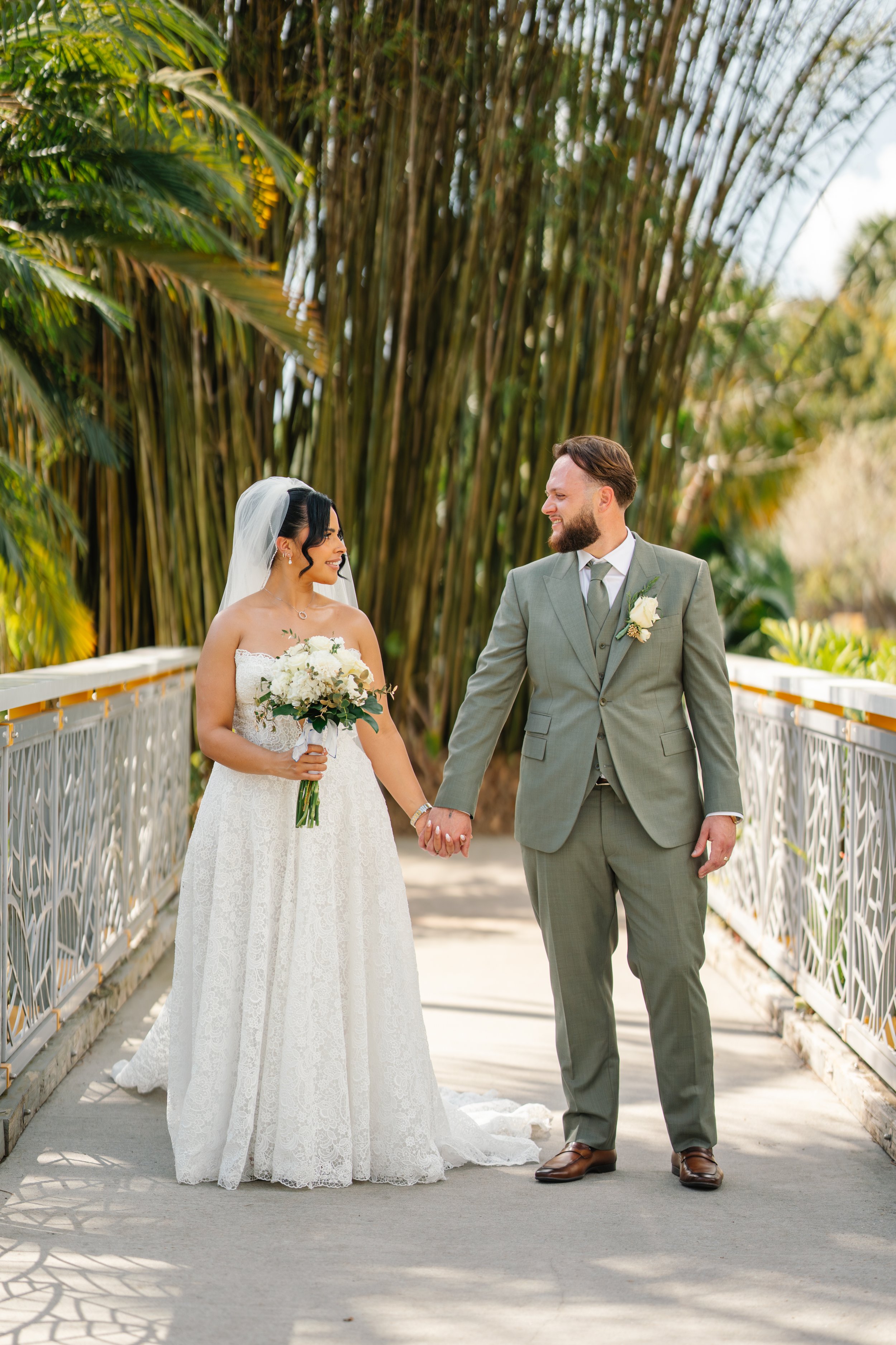 A bride and groom holding hands and smiling at each other on a walkway with greenery and tall trees in the background.