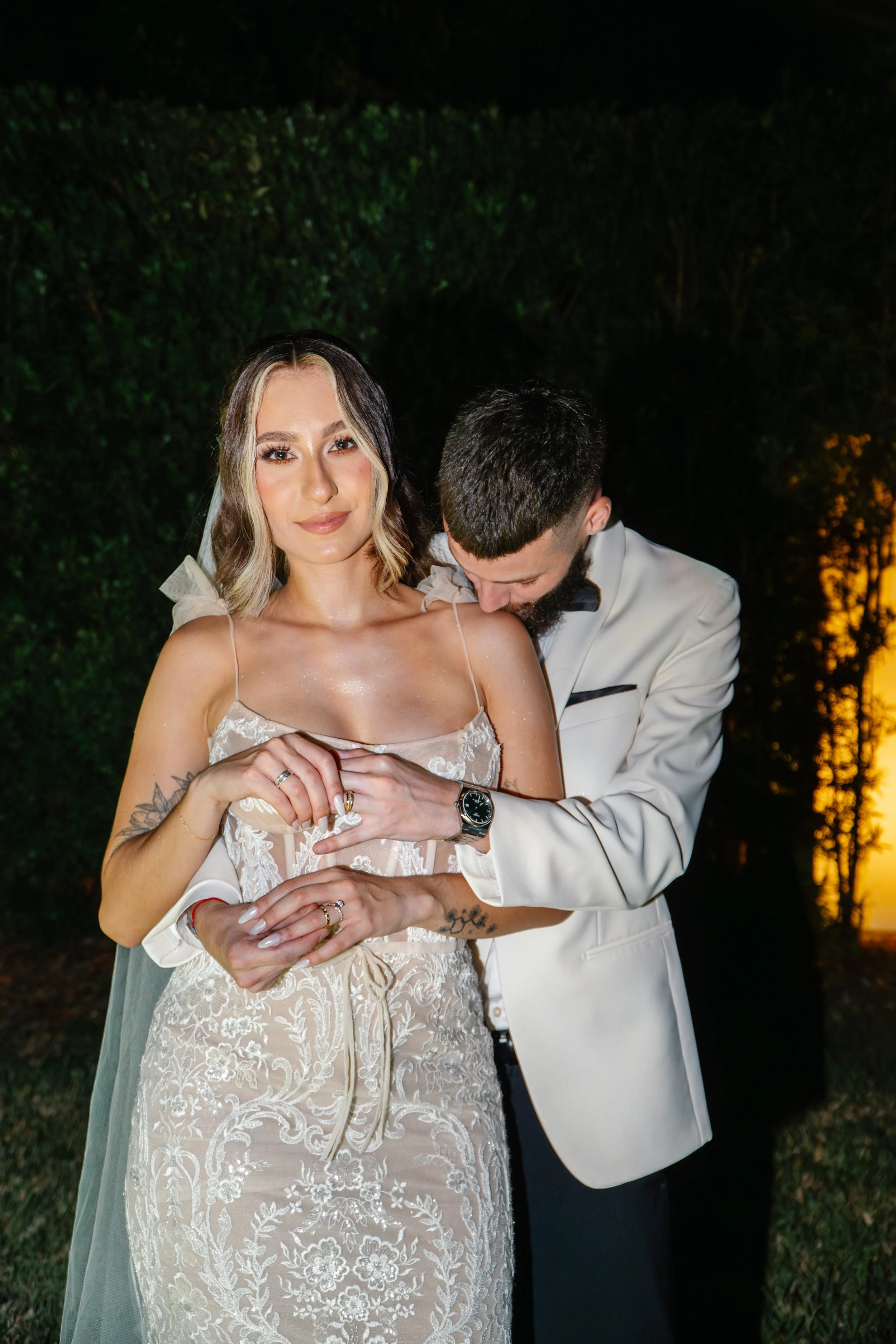 A bride and groom during their wedding, with the groom embracing the bride from behind and kissing her shoulder, standing outdoors at sunset.