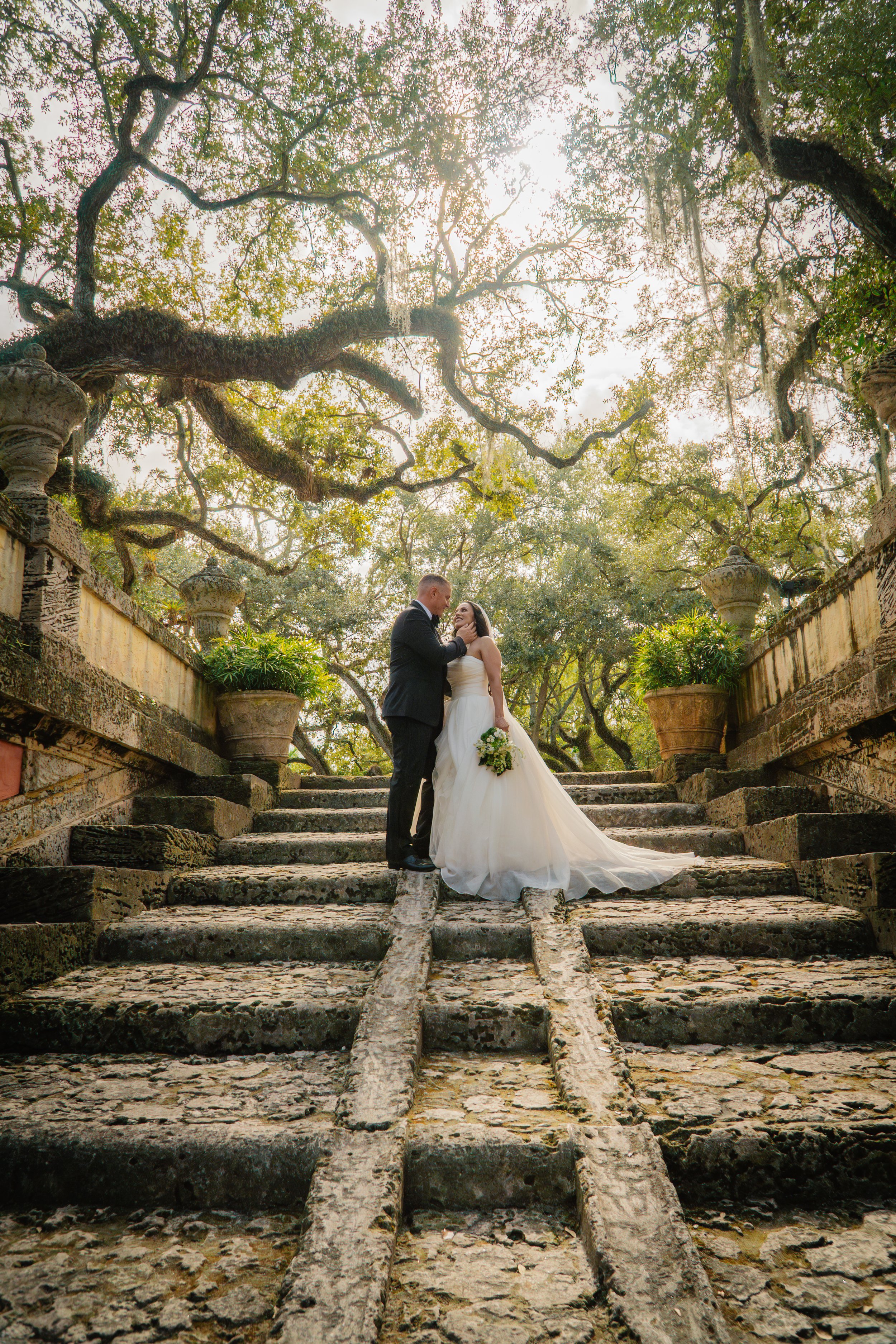 A bride and groom standing on moss-covered stone steps, surrounded by grand planters and large trees with hanging Spanish moss, with sunlight filtering through the branches.