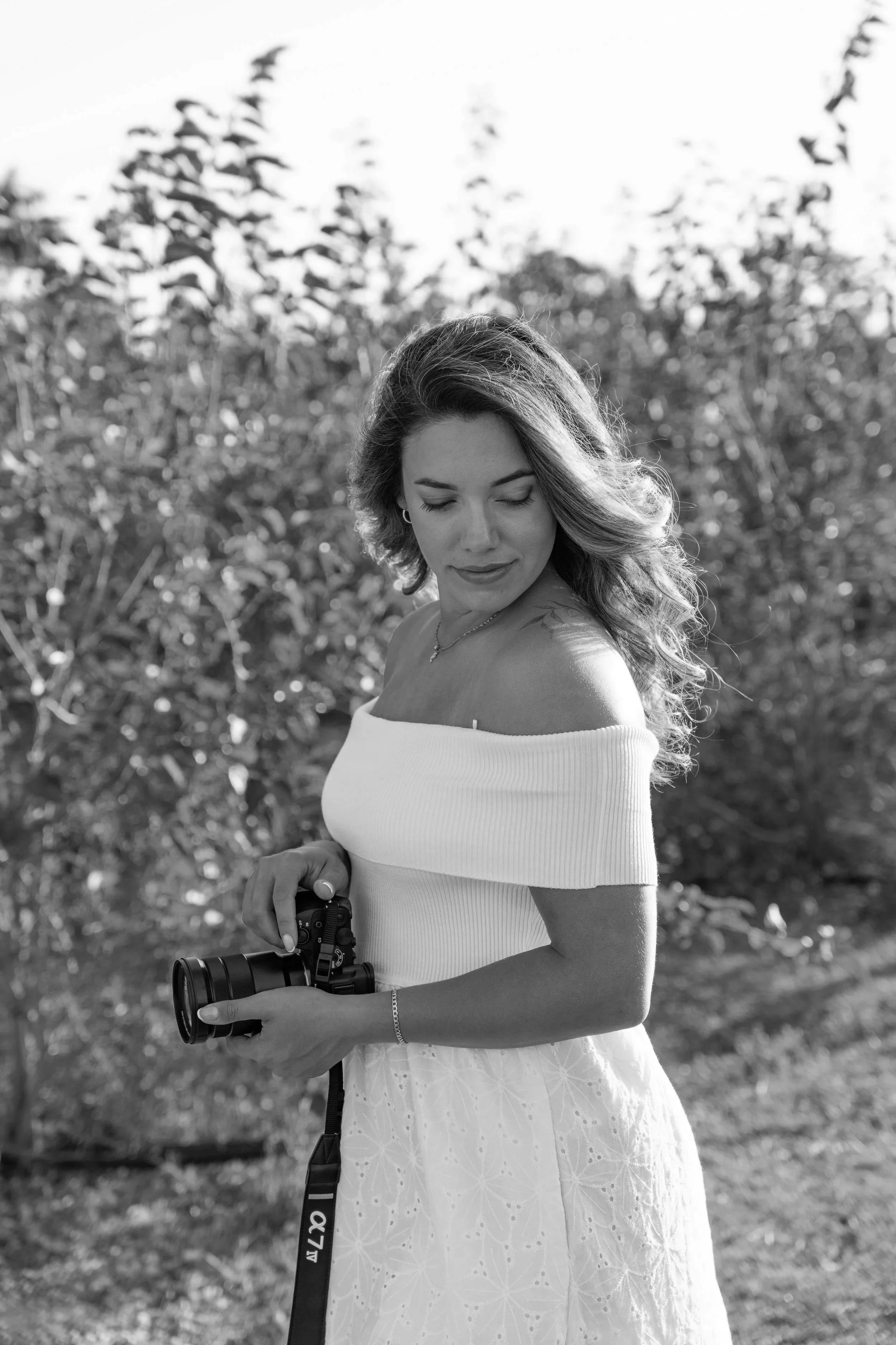 Black and white photo of a young woman outdoors, holding a camera, with trees in the background. She is wearing an off-shoulder top and a skirt, and has wavy hair.