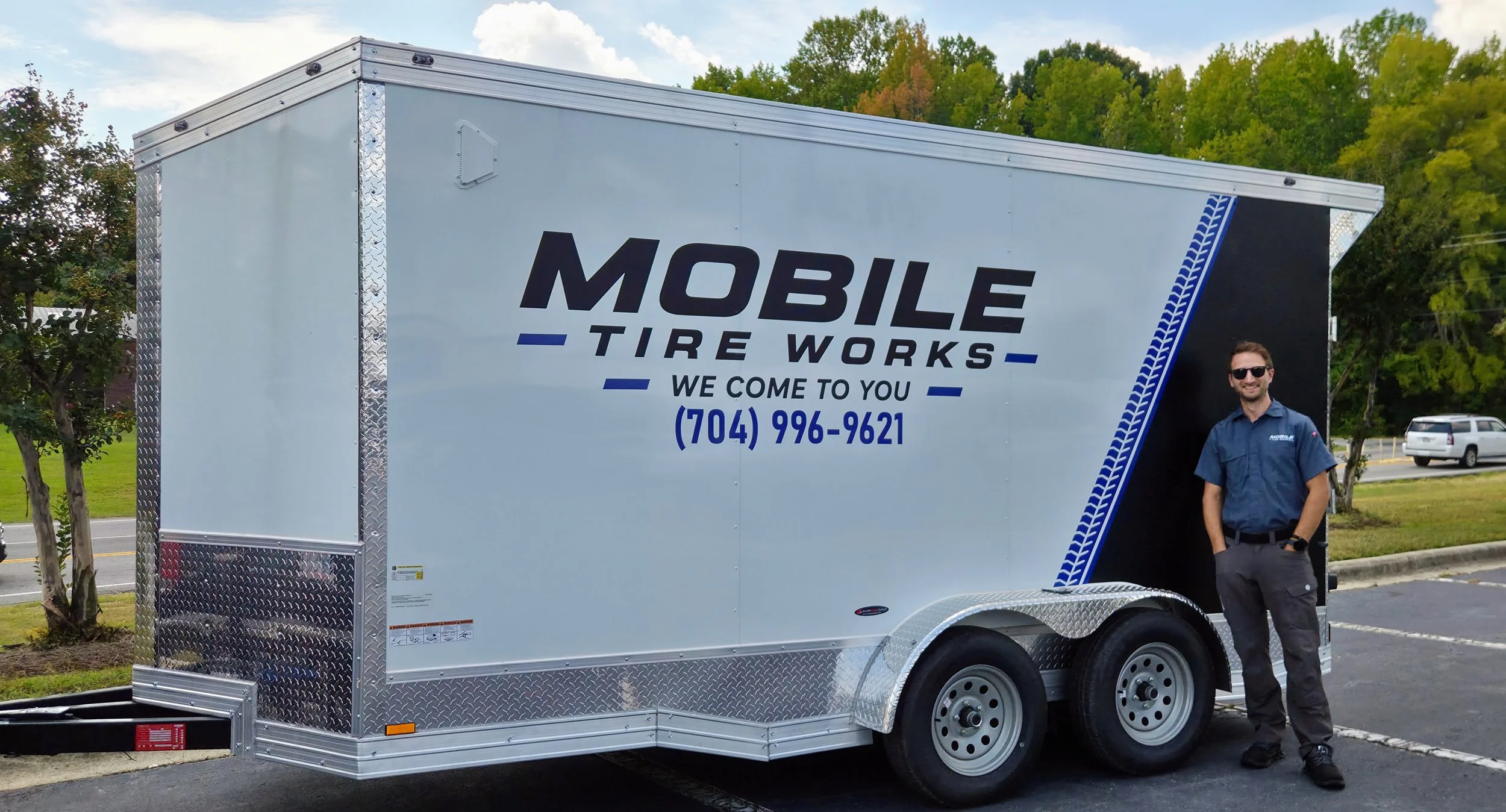 A mobile tire repair trailer with the text "Mobile Tire Works" and a phone number, parked outdoors with a smiling man in a uniform standing beside it.