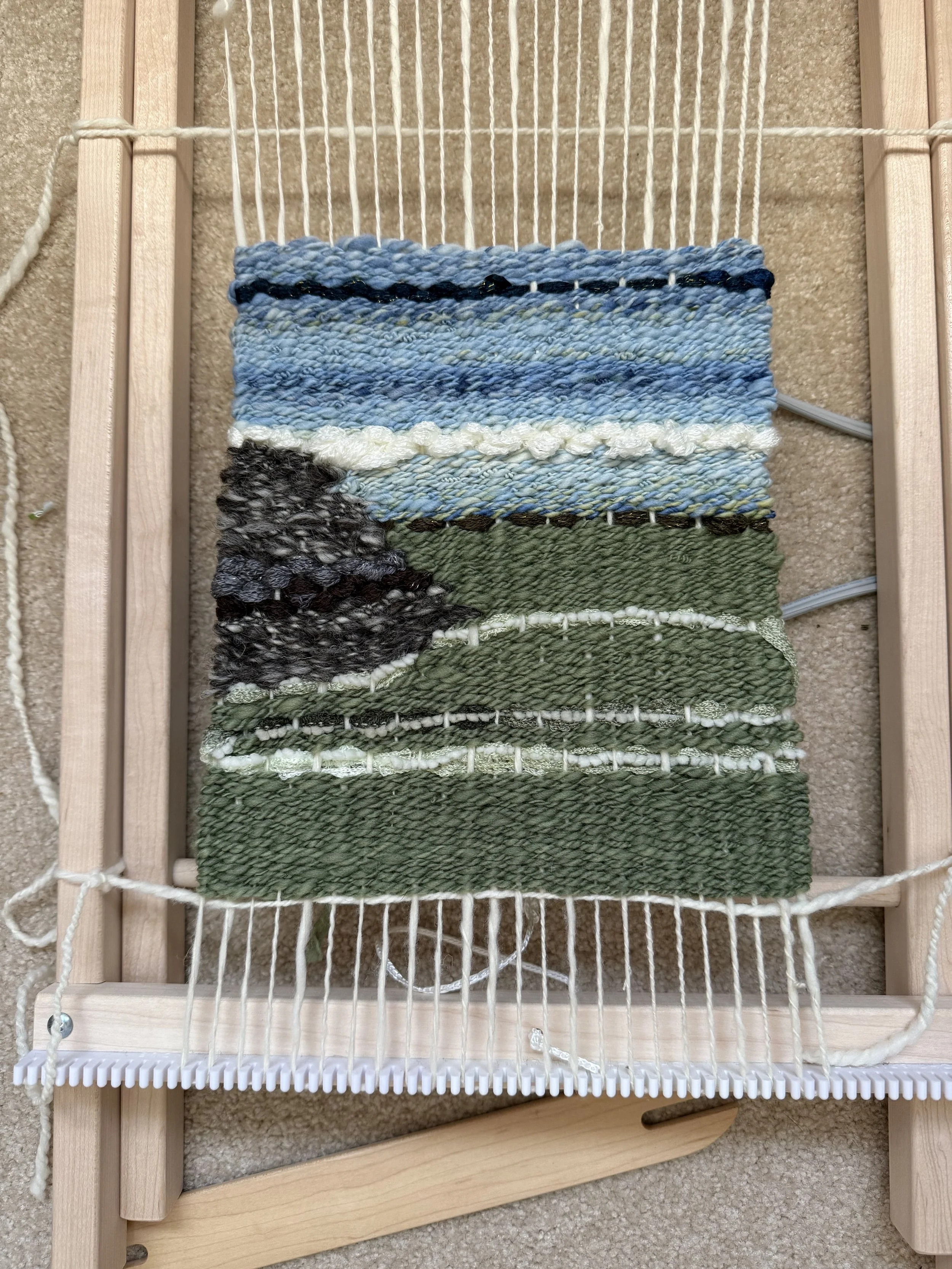 A frame loom with a woven image of a green field with fences and a rocky hill.