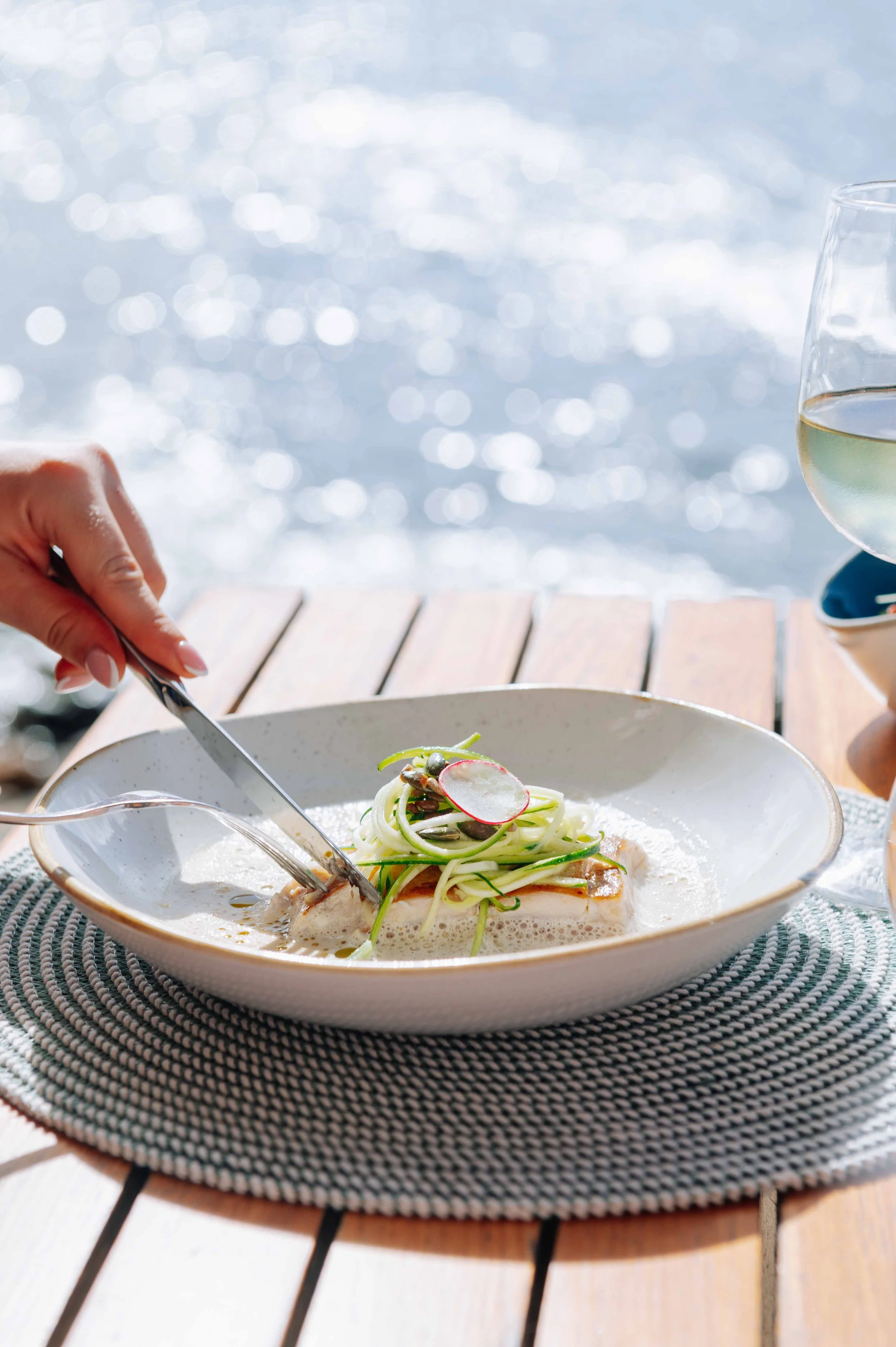 Assiette de poisson avec des légumes fins et radis, un verre de vin blanc, sur une table en bois près de l'eau en plein air.