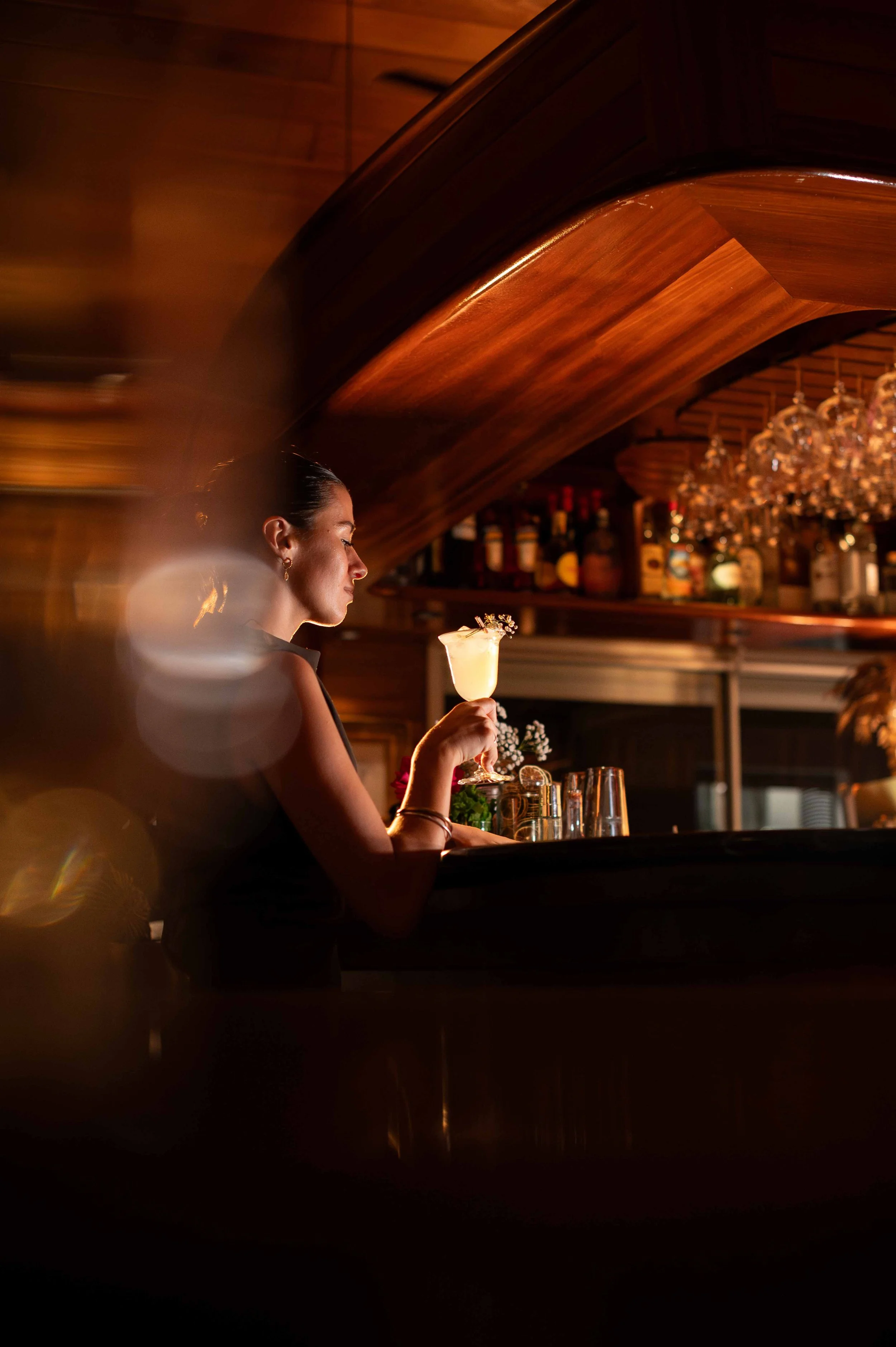 Une femme assise au bar dans un éclairage tamisé, tenant un cocktail dans un verre décoré, avec des bouteilles d'alcool en arrière-plan.