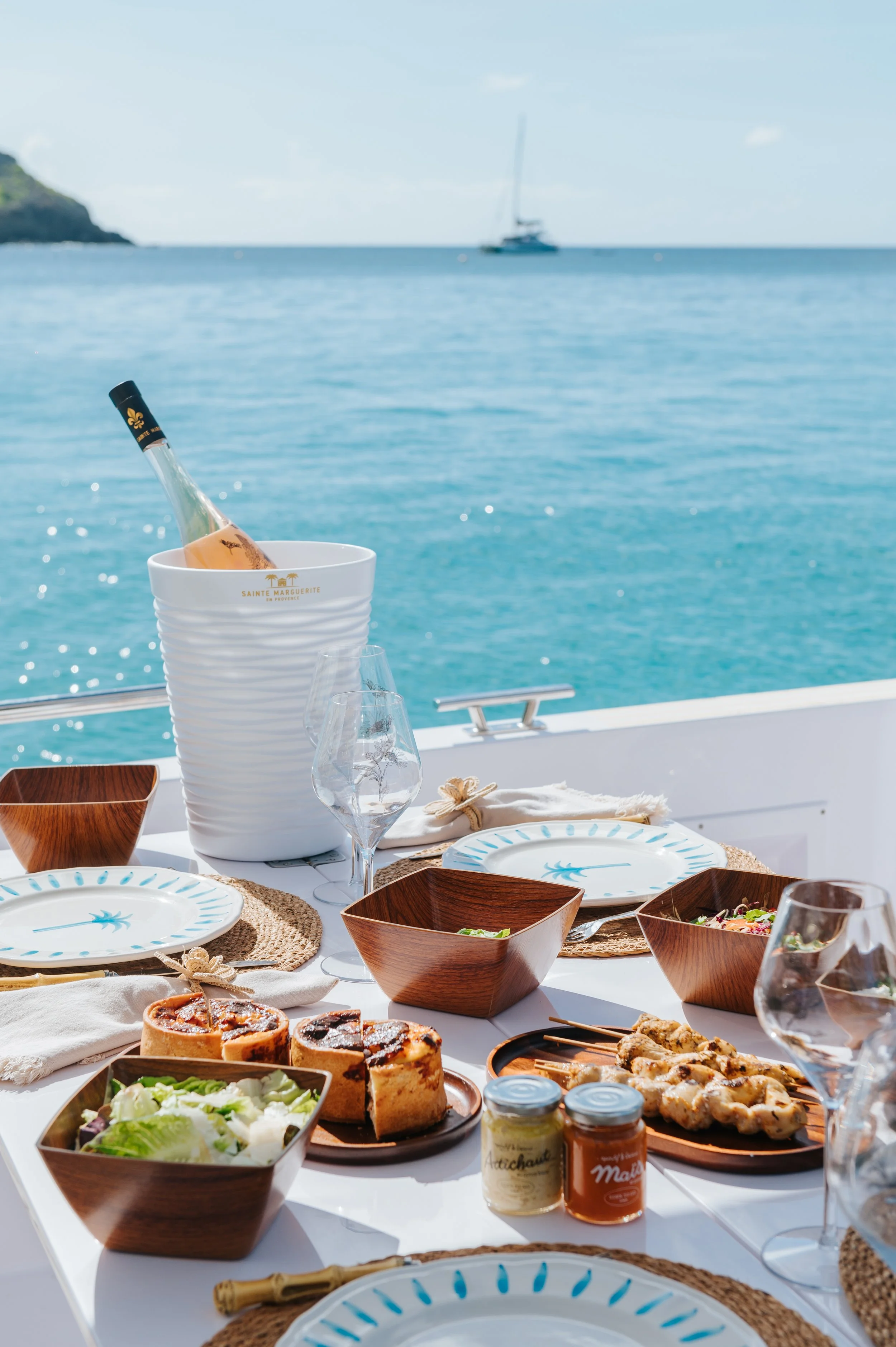 Repas de déjeuner en plein air avec des assiettes, des verres, des bocaux, une bouteille de champagne dans un seau en céramique, et une vue sur la mer avec un bateau au loin