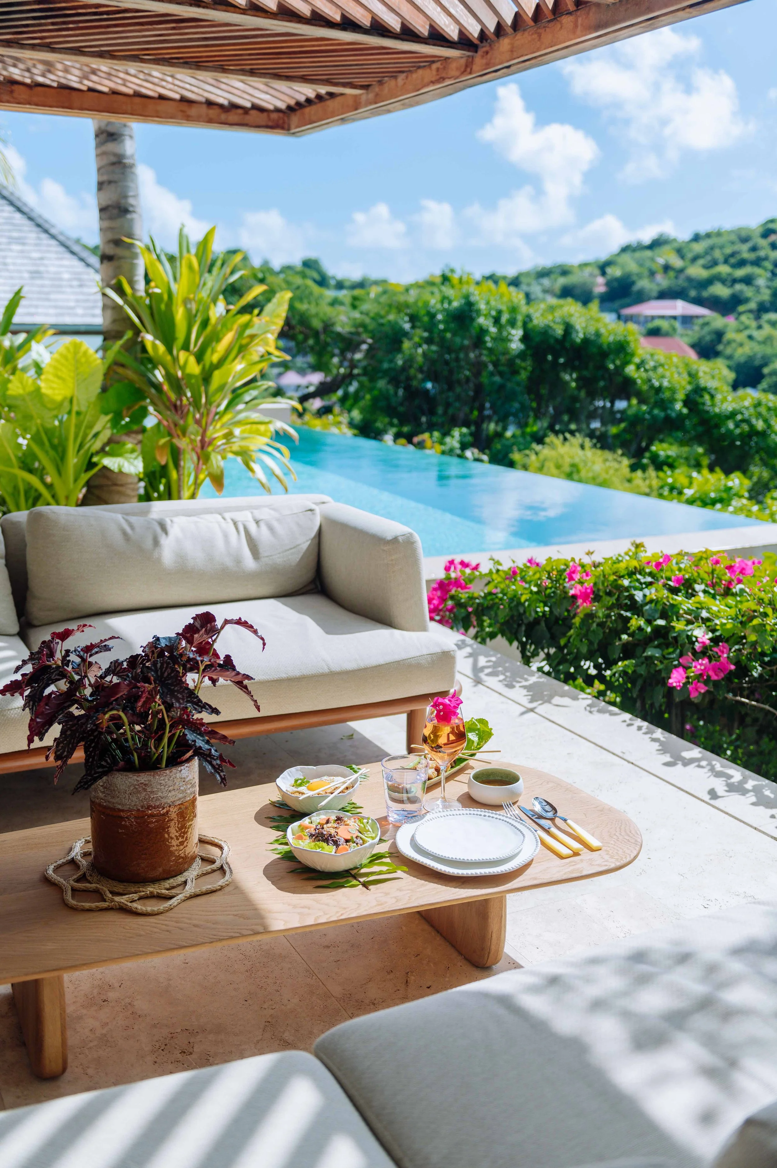 Un espace extérieur avec un canapé beige, une table en bois avec un repas, des plantes vertes, une piscine à débordement, un ciel bleu avec des nuages, et des fleurs roses.