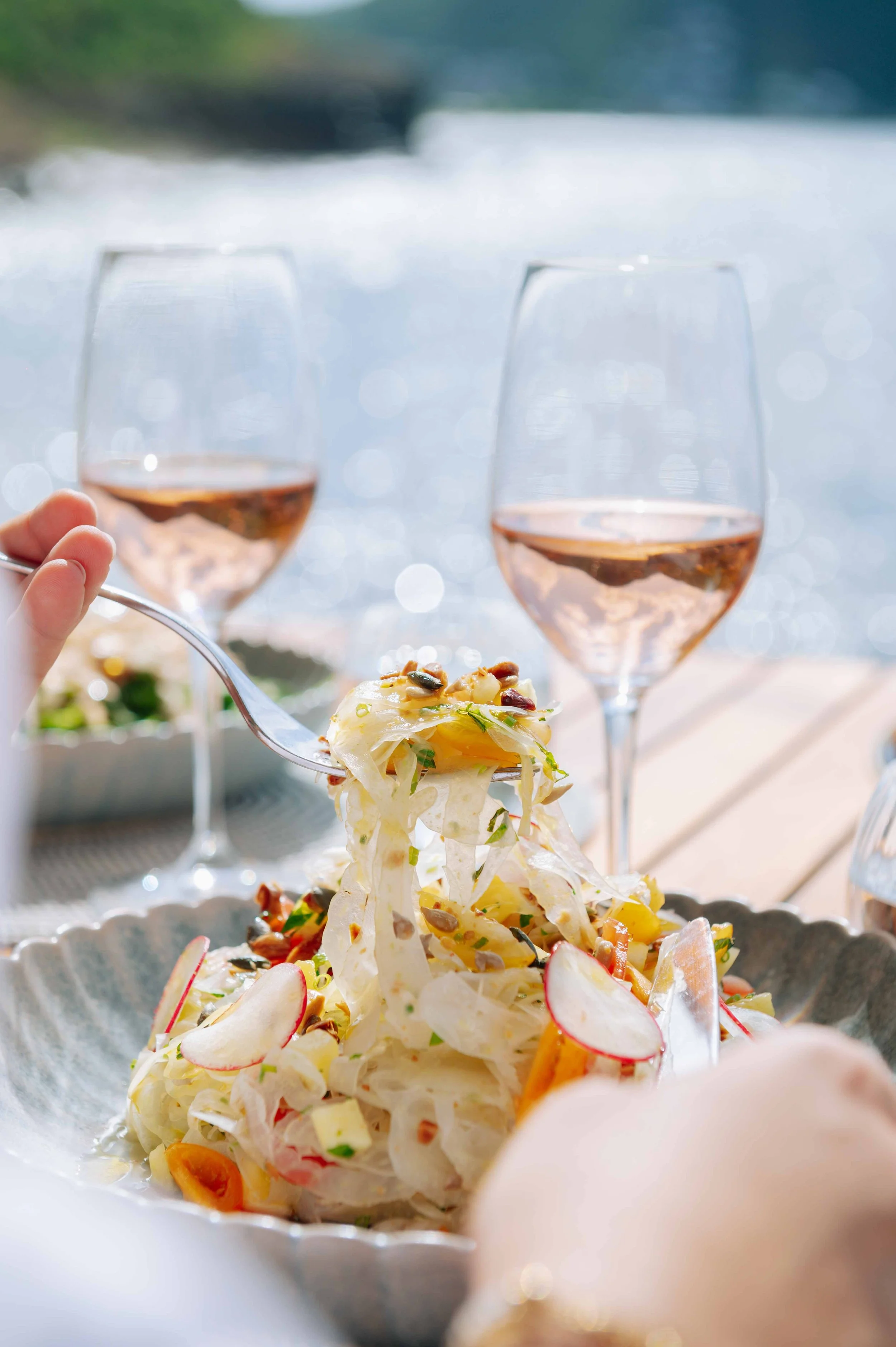 Un plat de salade avec des radis, des légumes, et des noix, accompagné de deux verres de rosé, en extérieur près d'un plan d'eau.