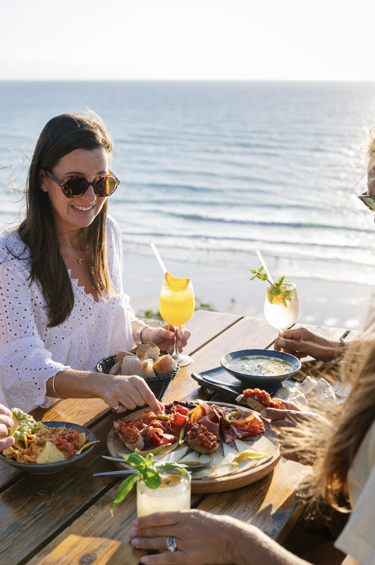 Group of women enjoying a meal and drinks at an outdoor seaside restaurant during sunset.
