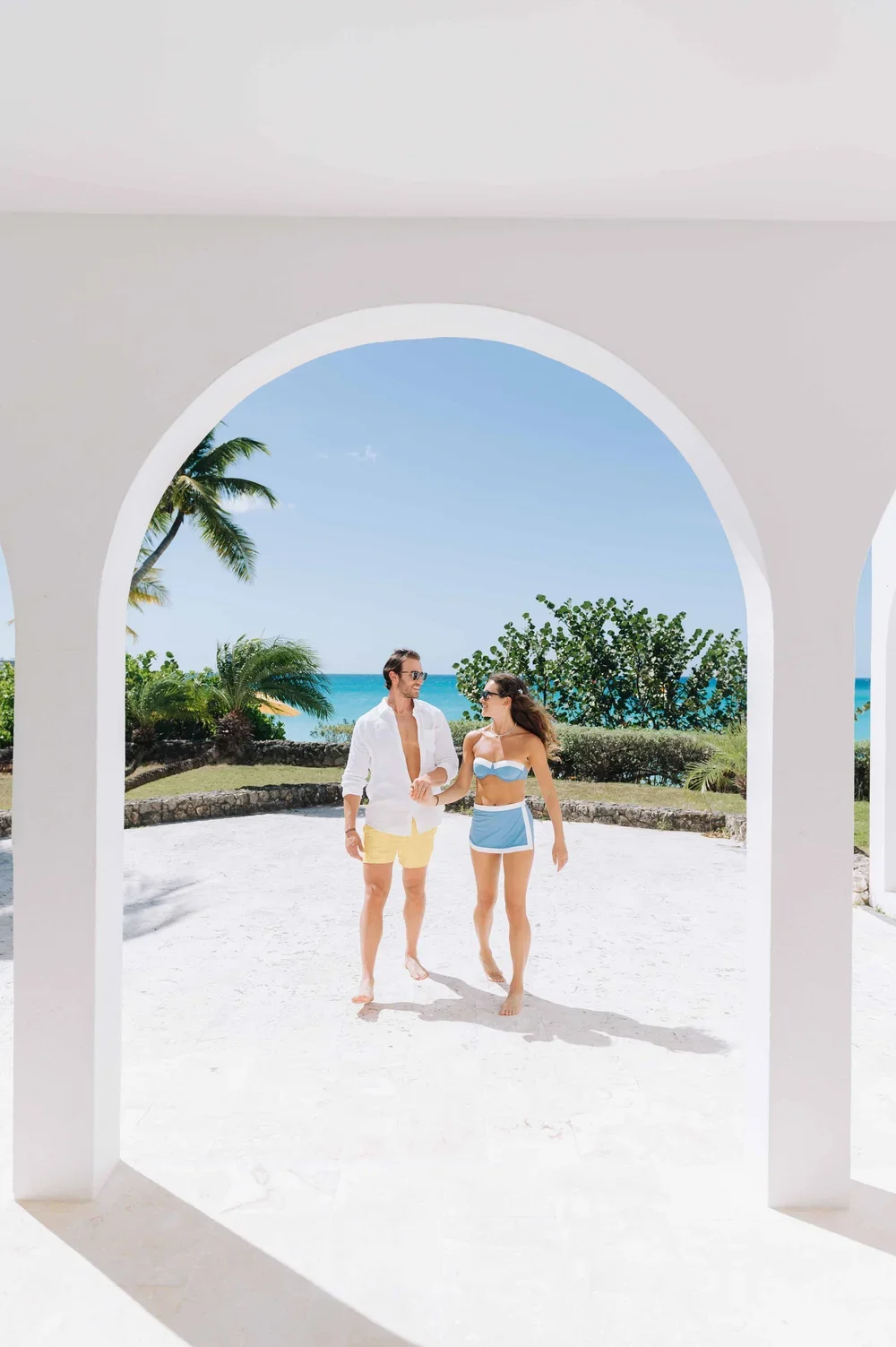 Un couple en maillot de bain marche sur une plage ensoleillée vue à travers une ouverture en forme d'arc dans un bâtiment blanc, avec des palmiers et la mer en arrière-plan.