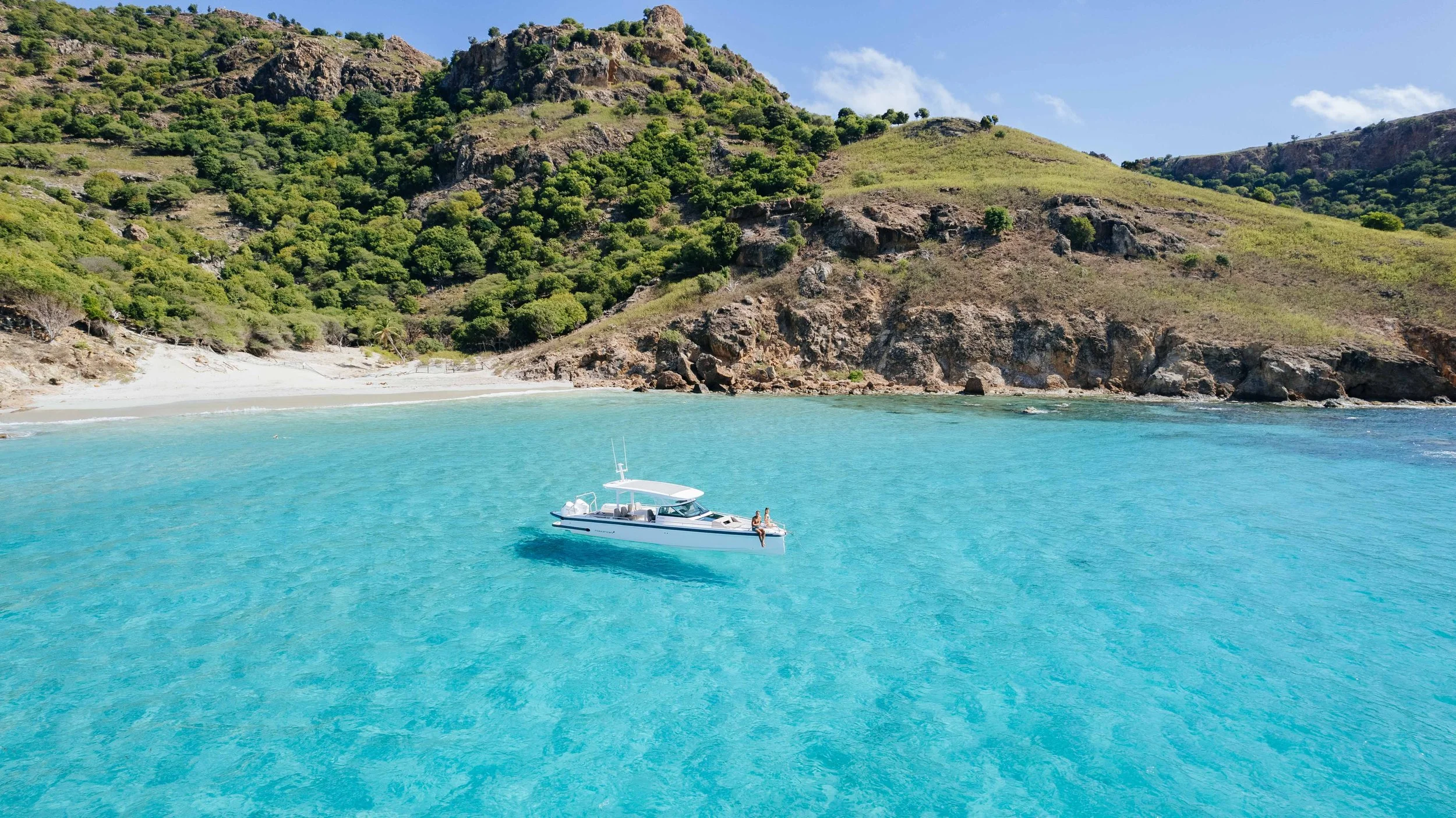 Une plage de sable blanc bordée de collines verdoyantes avec un bateau blanc flottant sur une mer turquoise calme.