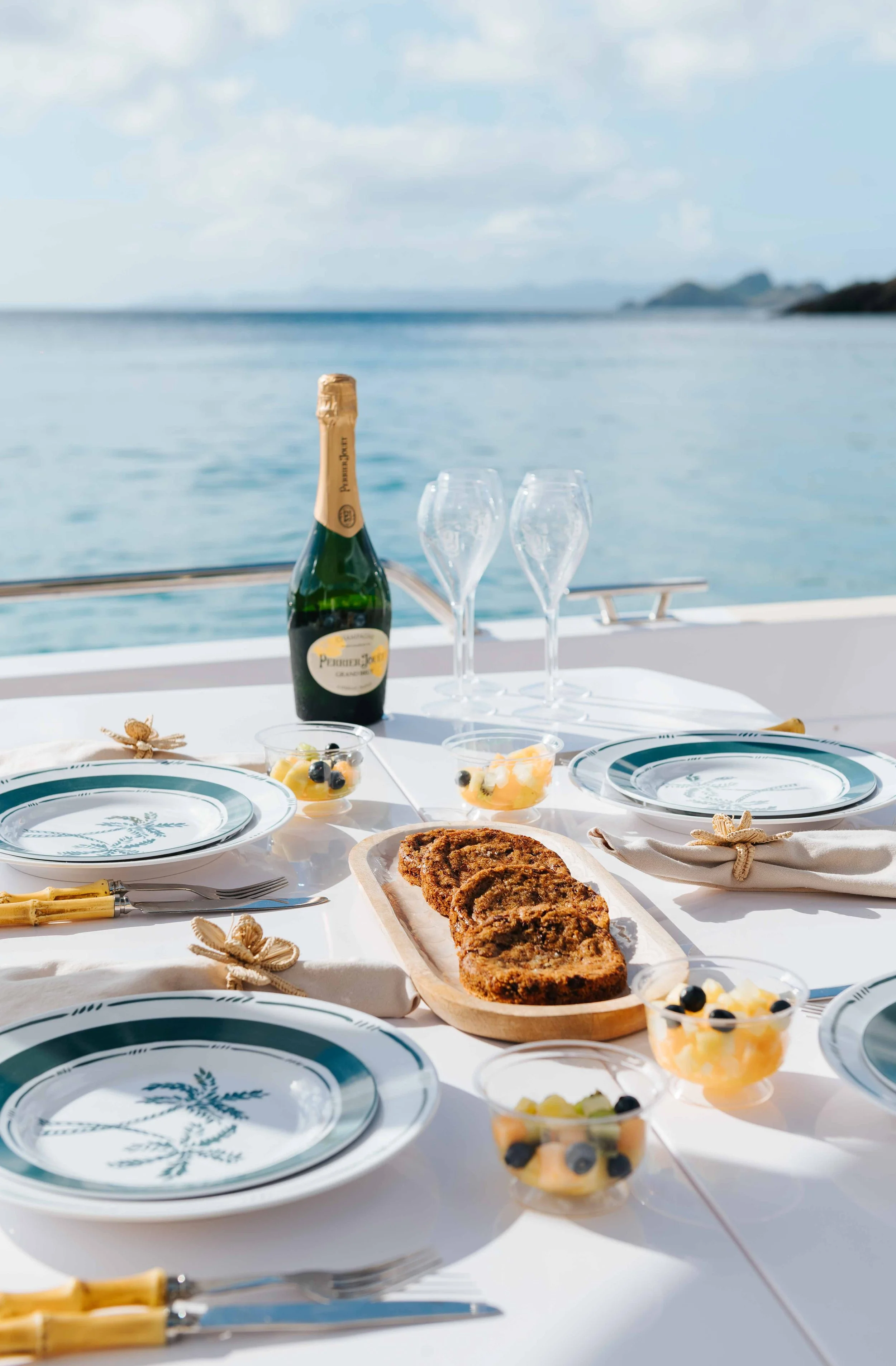 Table de repas en plein air avec une bouteille de champagne, deux flûtes, une planche de pain, des petits desserts et des couverts, avec la mer en arrière-plan.
