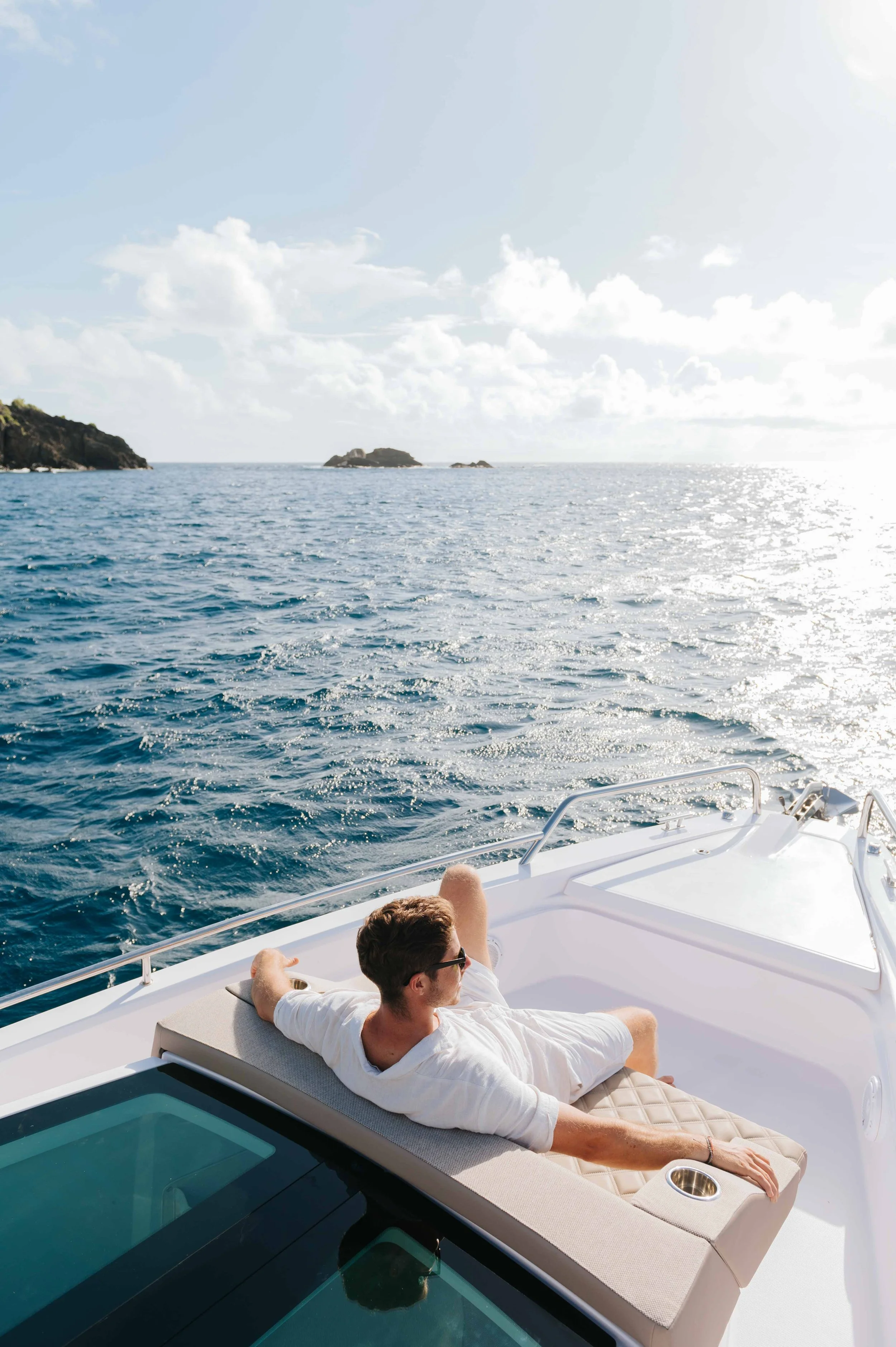 Un homme repose sur une chaise longue sur le pont d'un yacht, regardant l'océan avec de petites îles en arrière-plan, sous un ciel en partie nuageux.
